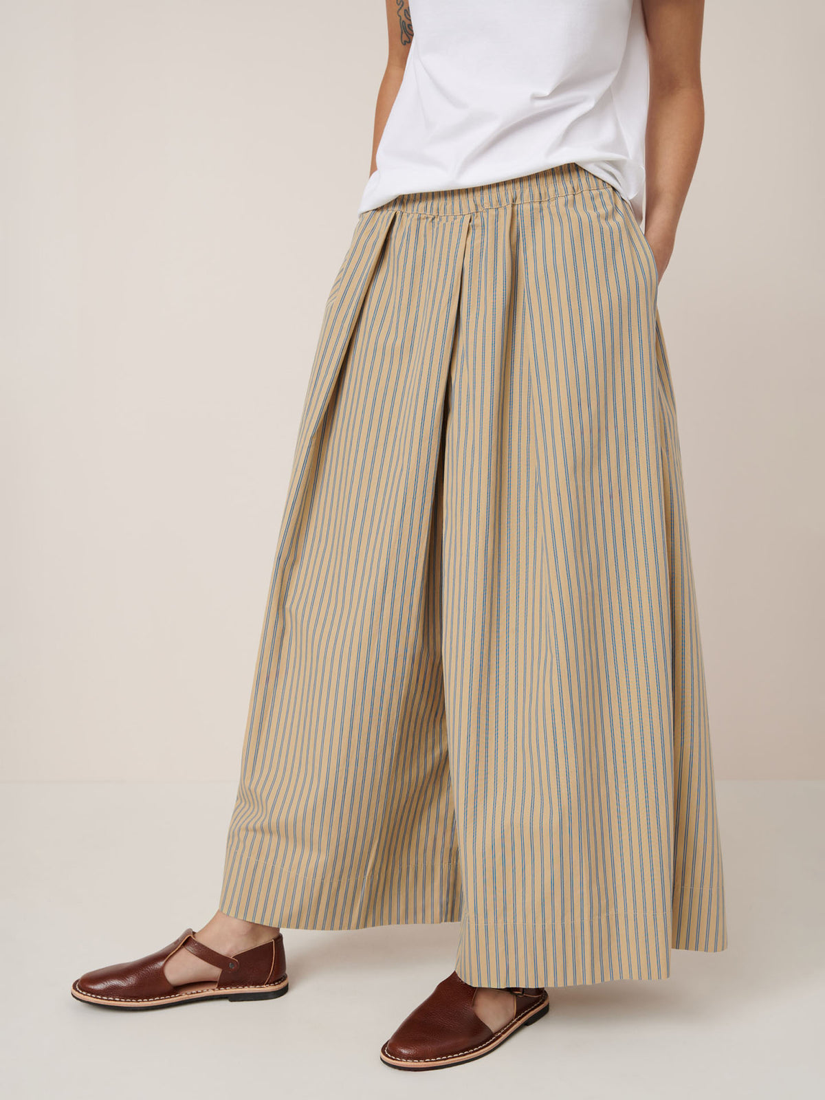 Person wearing Kowtow&#39;s Athena Pant – Double Pinstripe, featuring a relaxed fit, beige and blue vertical stripes, elastic waistband; paired with a white t-shirt and brown flats, hand in pocket.