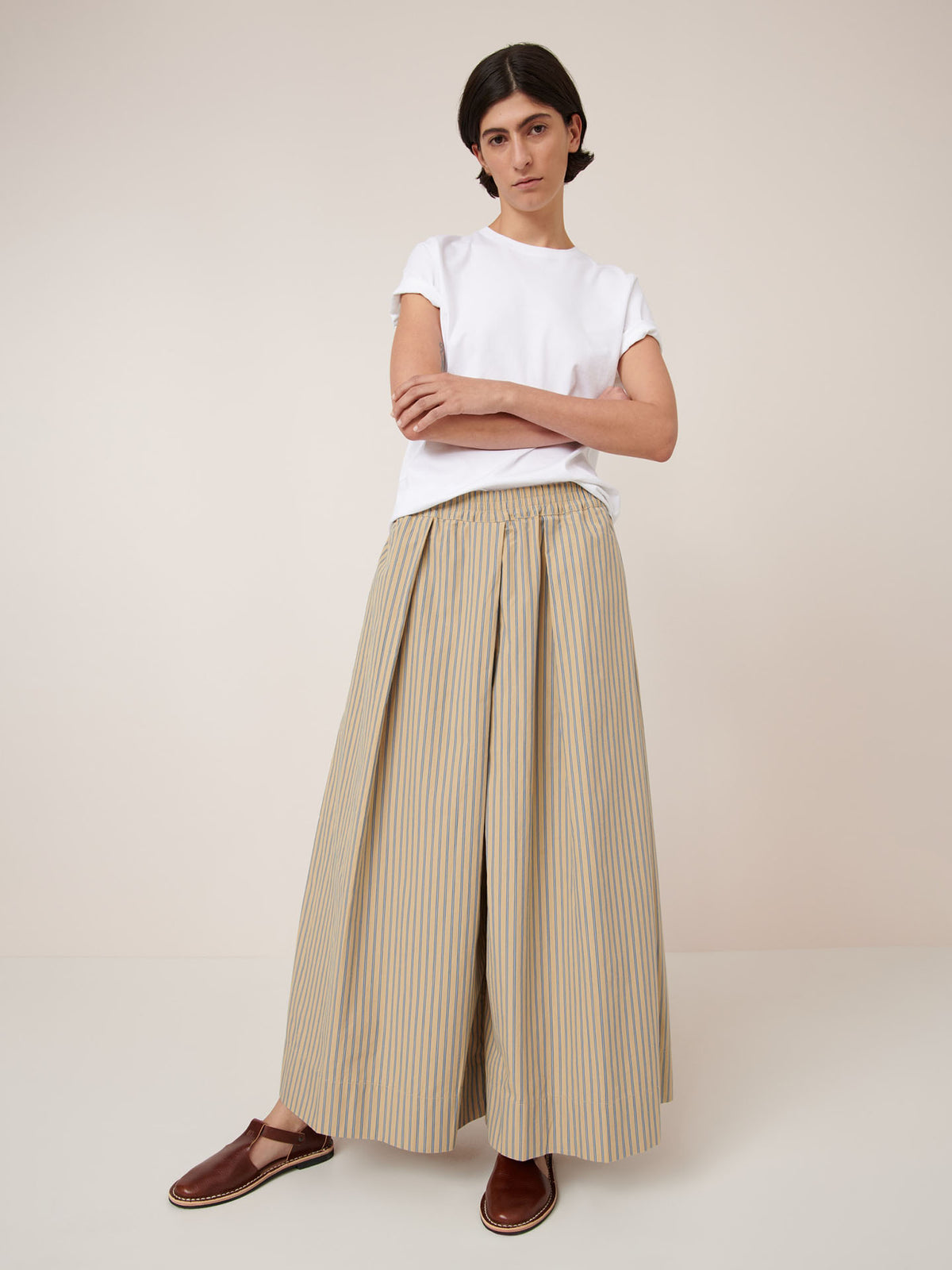 A person stands with arms crossed, wearing a white t-shirt and Kowtow&#39;s Athena Pant – Double Pinstripe, featuring a true waist and elastic waistband in beige and blue stripes, paired with brown flat shoes, against a light background.