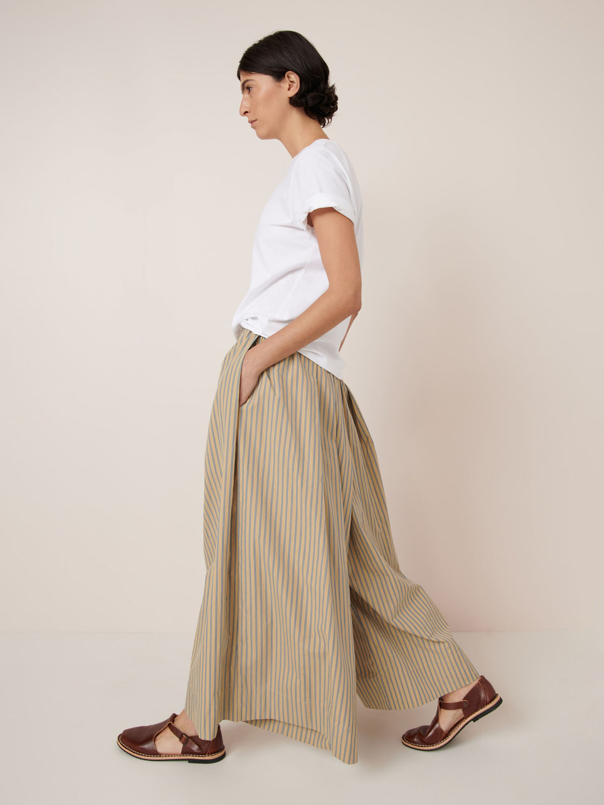 A person in profile wears Kowtow’s Athena Pant – Double Pinstripe, featuring beige and yellow stripes with an elastic waistband, styled with a white t-shirt and brown sandals against a plain light background.