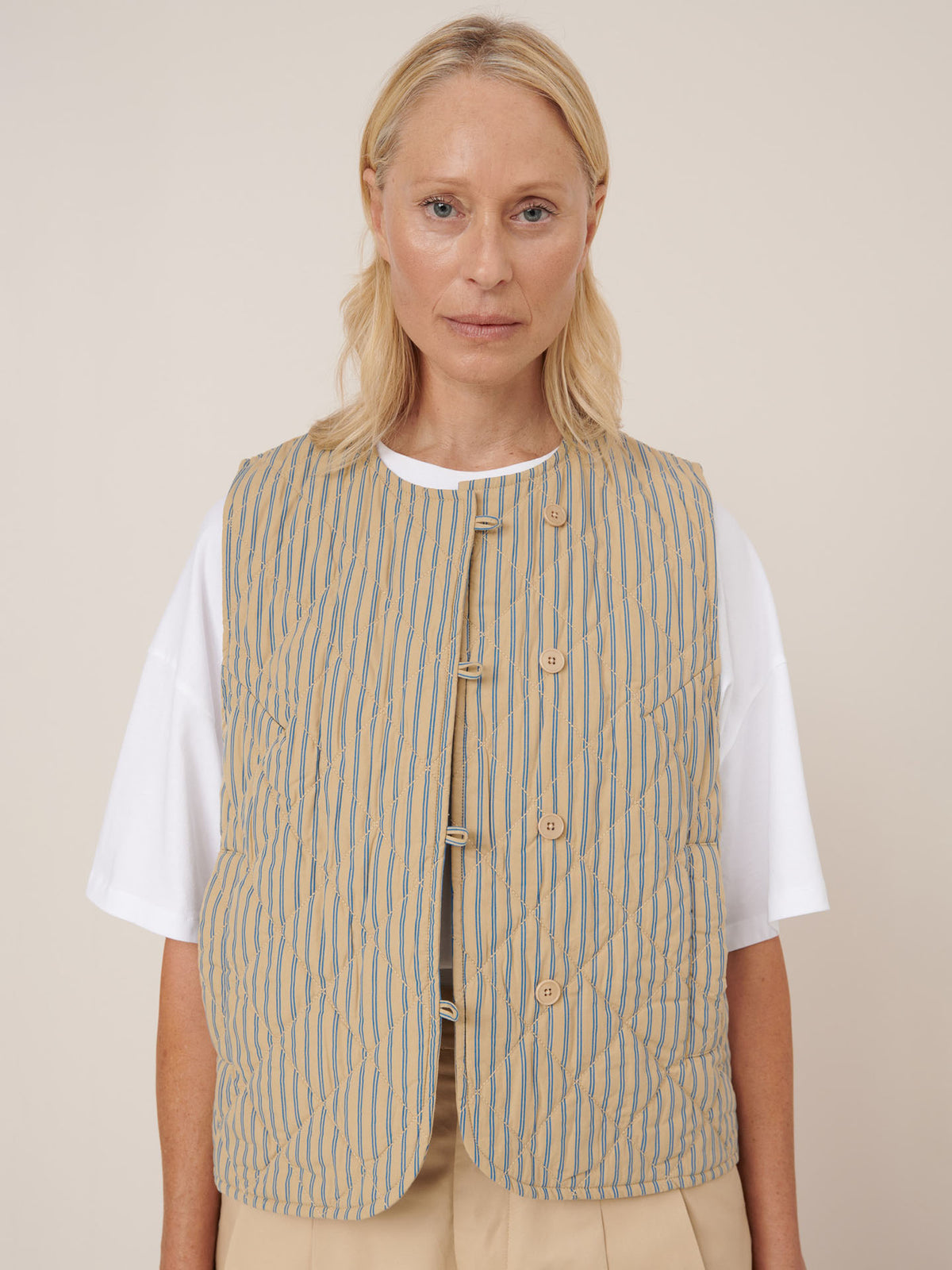 A woman with blonde hair wears the Kowtow Axis Vest – Double Pinstripe, featuring beige and blue stripes, over a white T-shirt against a plain light background. The vest provides a standard fit and is true to size for effortless styling.