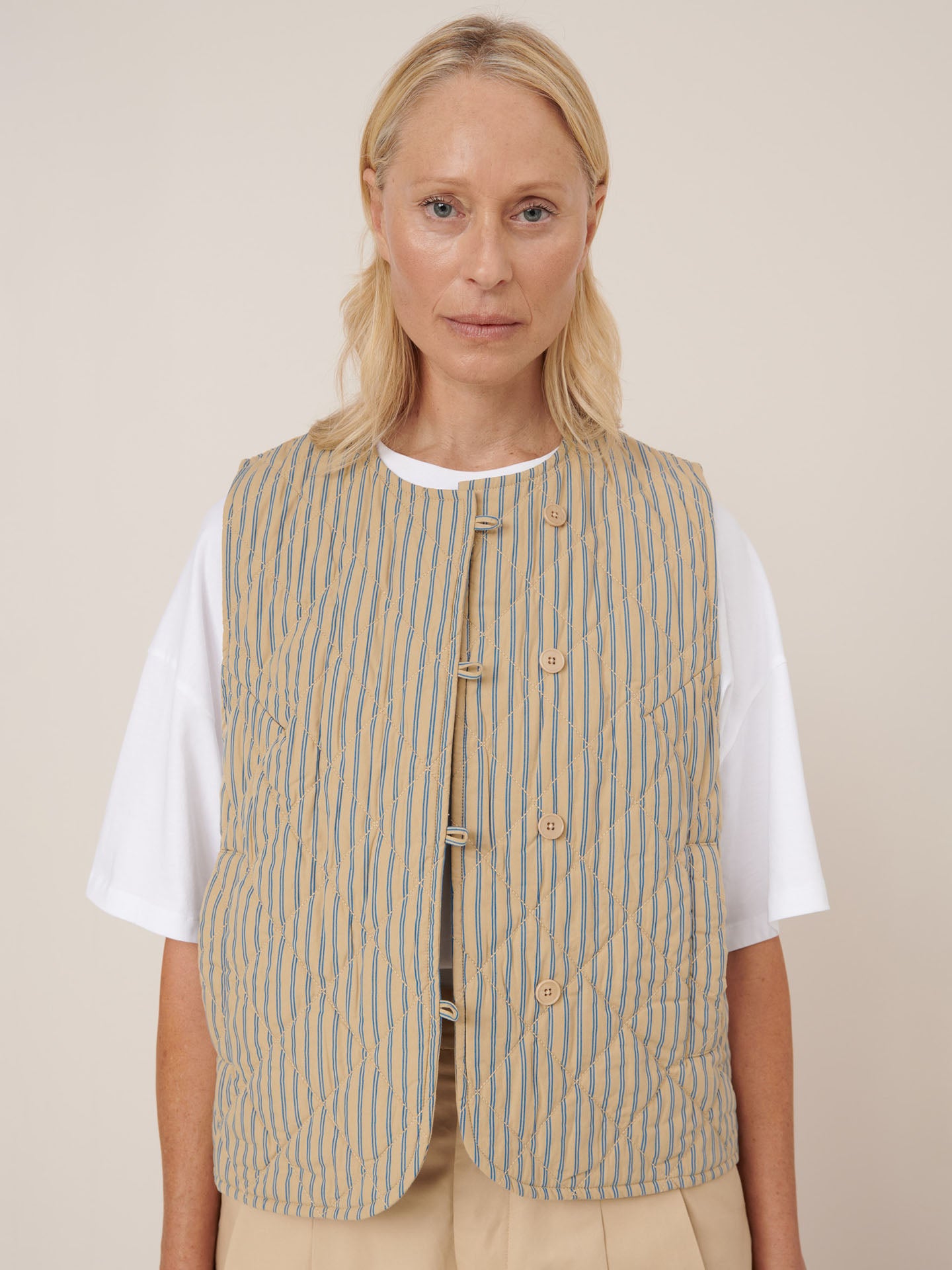 A woman with blonde hair wears the Kowtow Axis Vest – Double Pinstripe, featuring beige and blue stripes, over a white T-shirt against a plain light background. The vest provides a standard fit and is true to size for effortless styling.