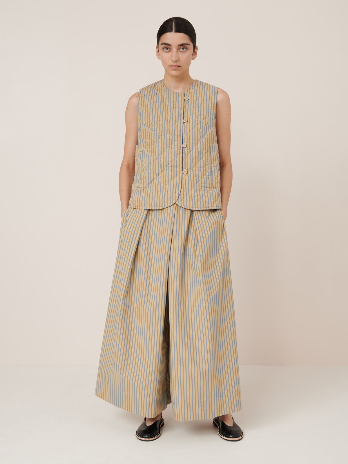 A person stands against a plain background wearing the Kowtow Axis Vest – Double Pinstripe and matching wide-legged trousers, both true to size, with hands in pockets and black slip-on shoes.