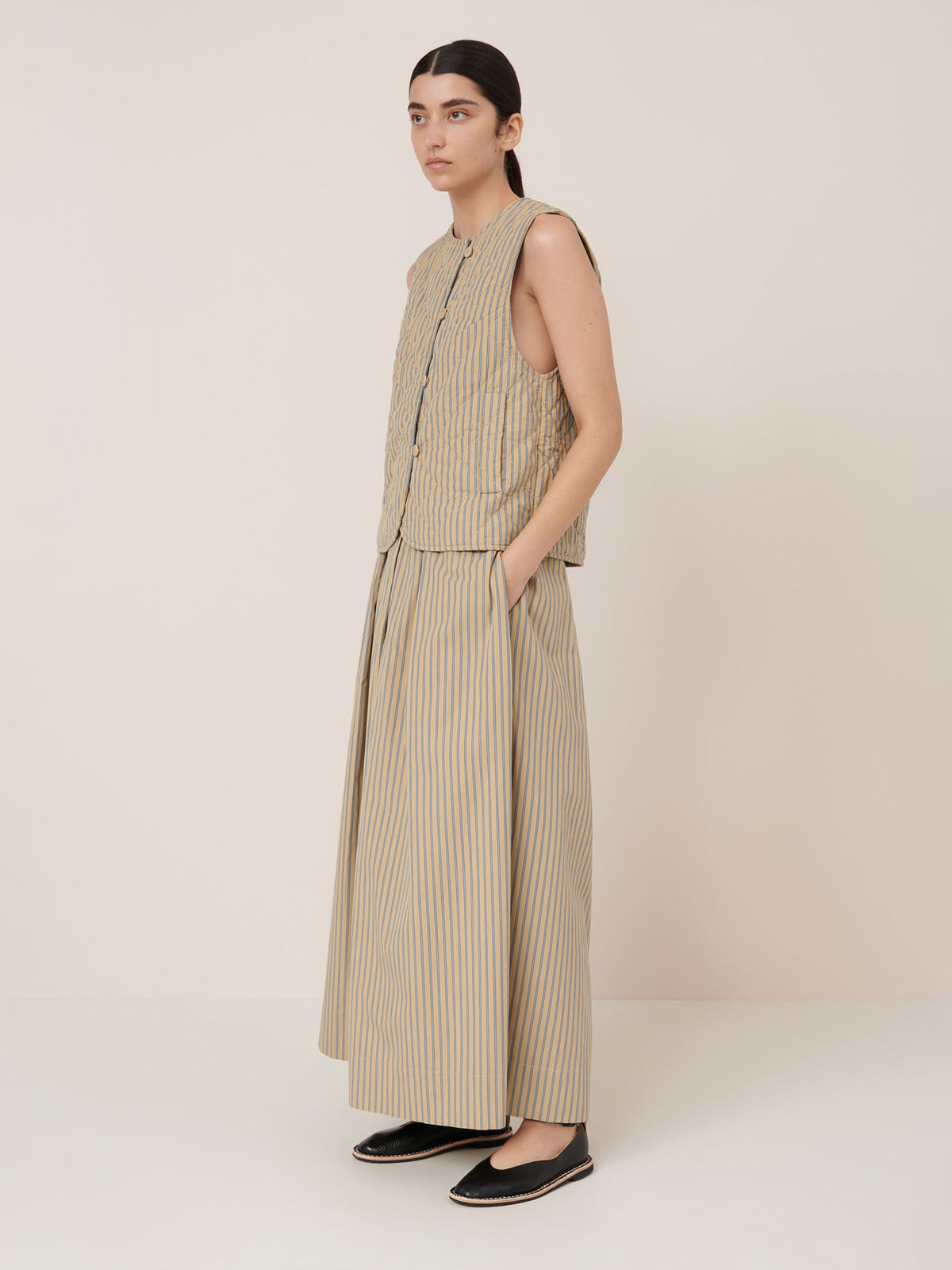 A woman stands against a plain background, wearing the Kowtow Axis Vest – Double Pinstripe, a sleeveless quilted buttoned vest with pockets, paired with a long striped skirt and black slip-on shoes for a true-to-size fit.