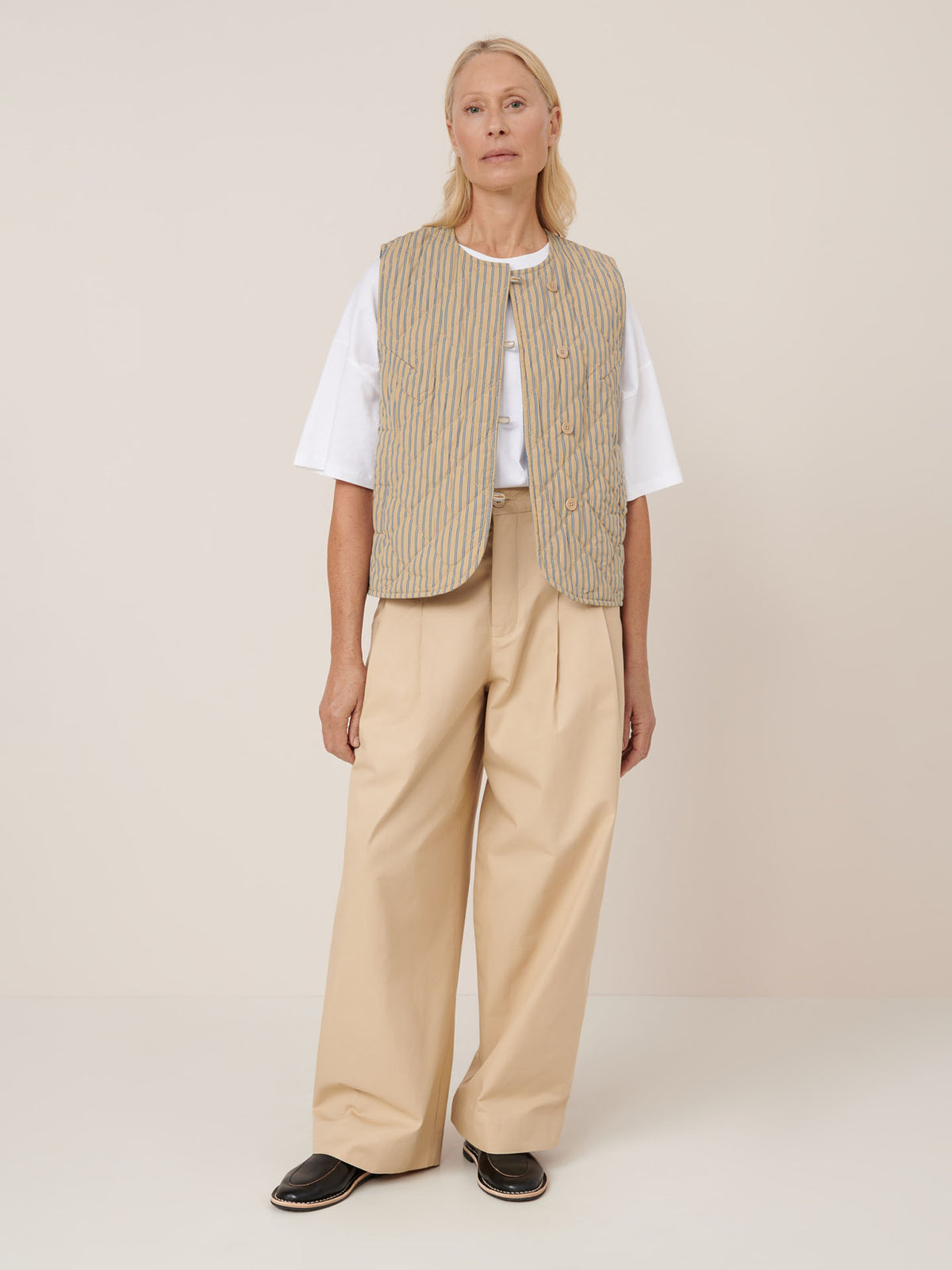 A person stands against a plain background wearing the Kowtow Axis Vest – Double Pinstripe over a white t-shirt, plus beige wide-leg trousers and black slip-on shoes, showcasing its standard fit that&#39;s true to size for accurate measurements.