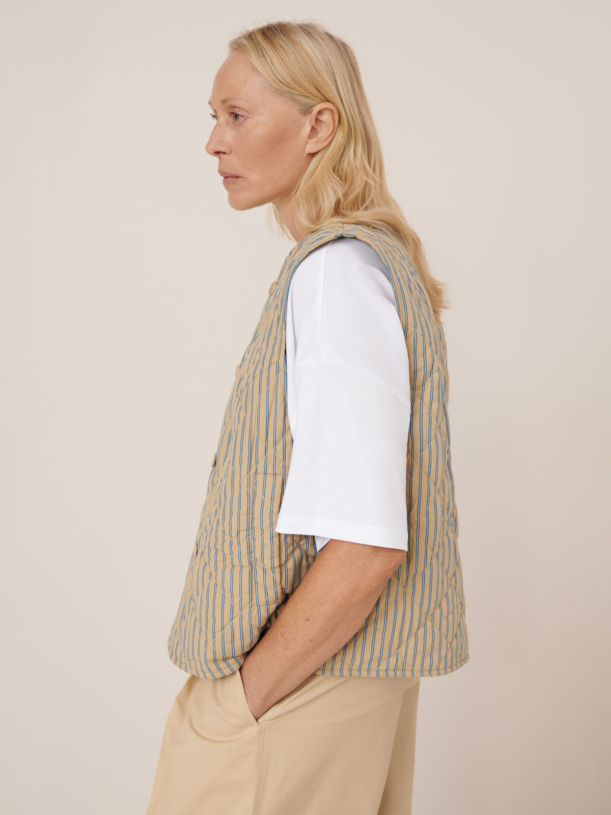 A person with long blonde hair stands in profile with one hand in their pocket, wearing a white t-shirt, beige pants in a standard fit, and the Kowtow Axis Vest – Double Pinstripe (striped beige and blue vest).
