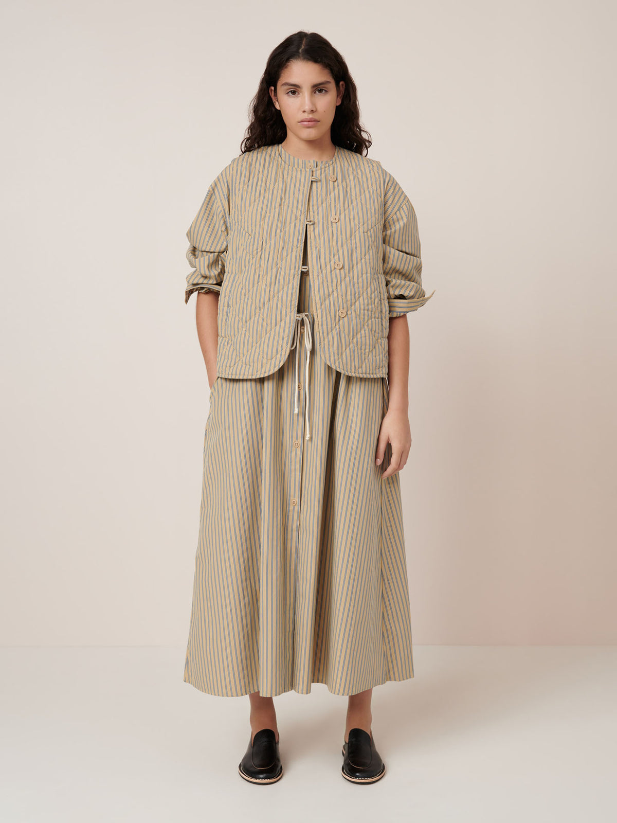 A person wears the Kowtow Axis Vest – Double Pinstripe against a plain background with black slip-on shoes, looking at the camera. The vest features standard fit garment measurements and is true to size.