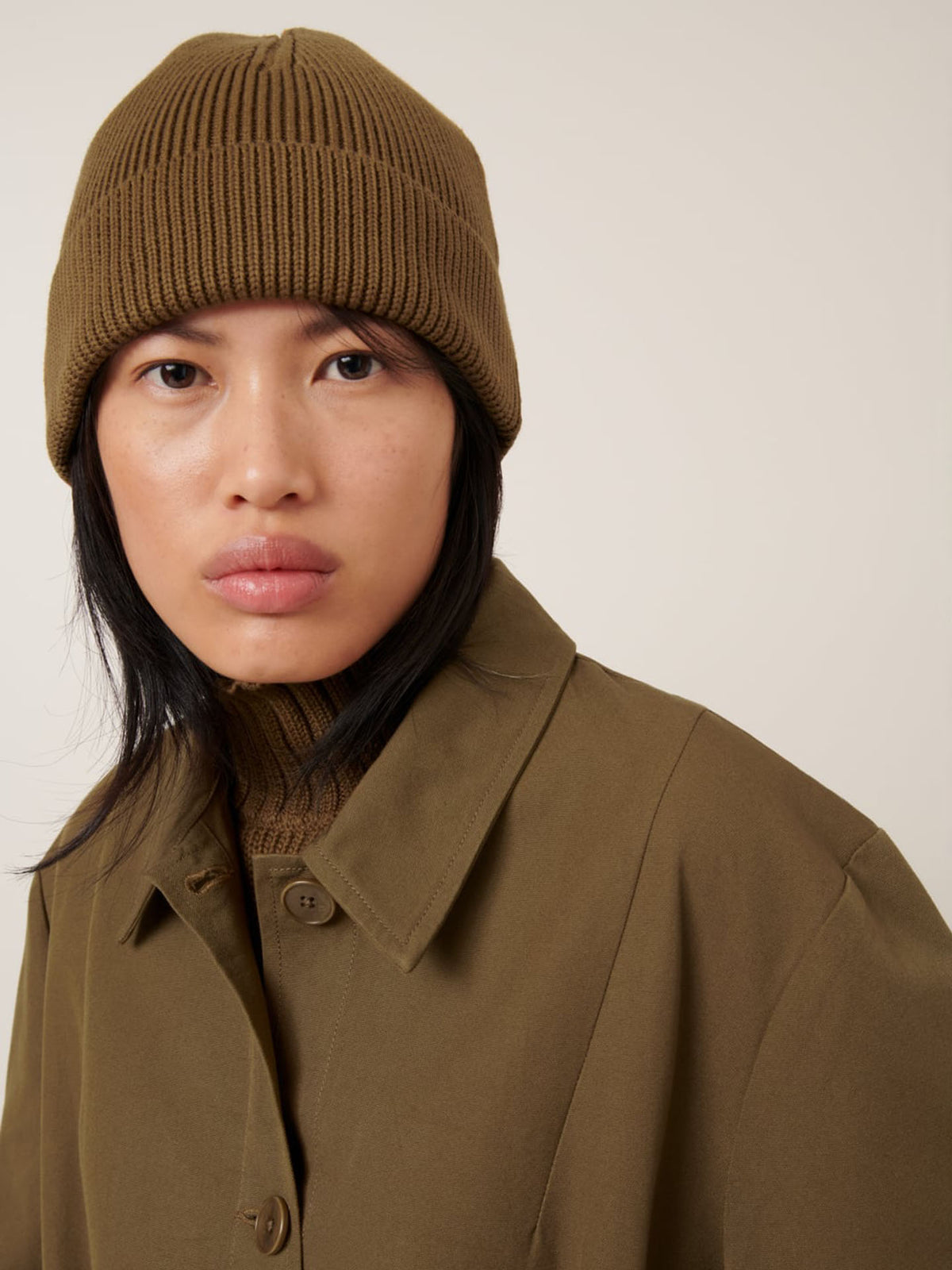 A person wearing the Kowtow Beanie – Dark Spice, paired with a brown turtleneck and coat, looks at the camera against a plain light background.