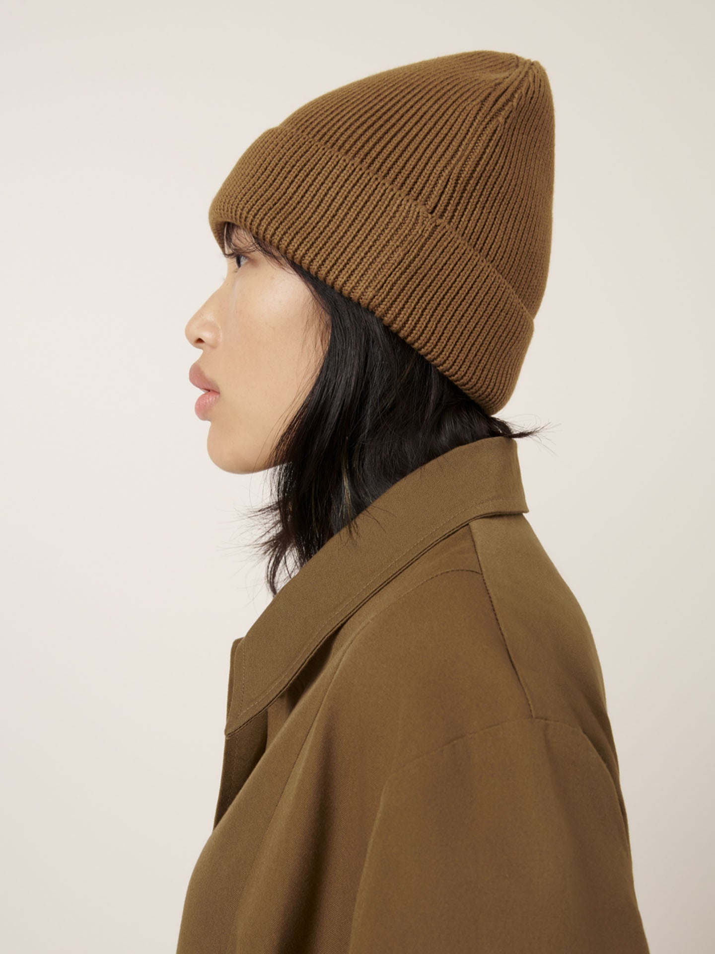 A person with long black hair is shown in profile against a plain background, wearing Kowtow's Beanie – Dark Spice and a brown coat.