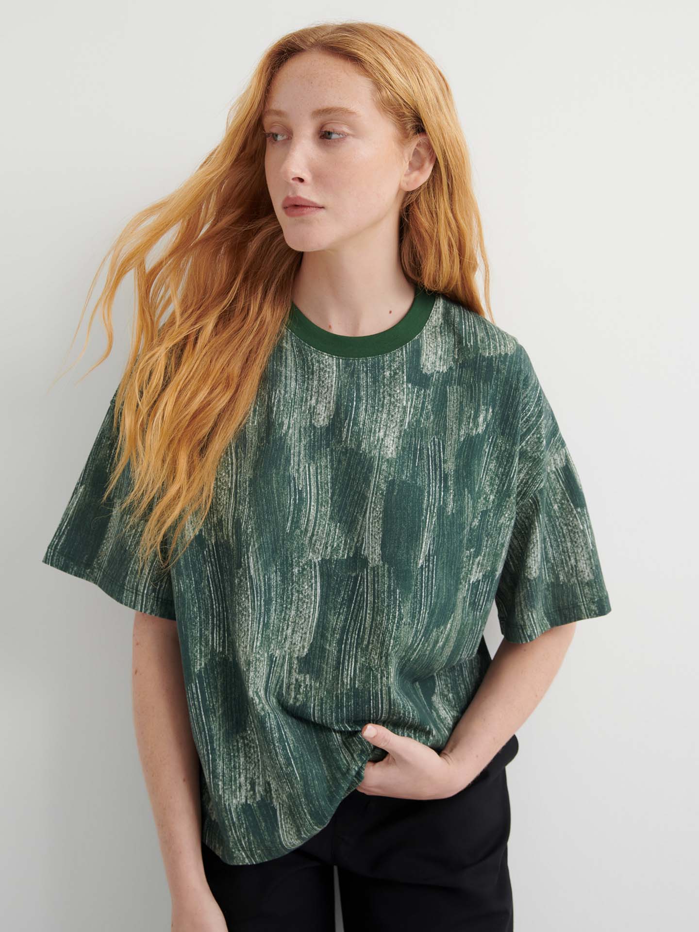 A person with long red hair wears the Brushstroke Tee, an oversized boxy top with a green pattern, paired with black pants, standing against a plain light background.