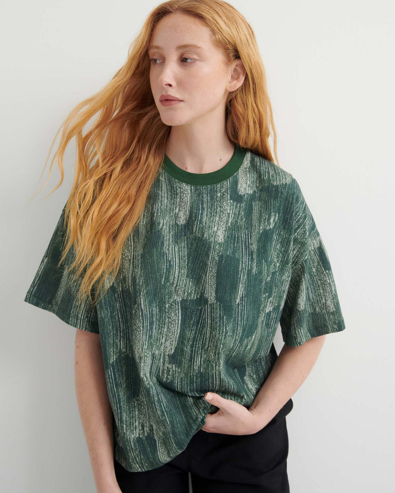 A person with long red hair wears the Brushstroke Tee, an oversized boxy top with a green pattern, paired with black pants, standing against a plain light background.