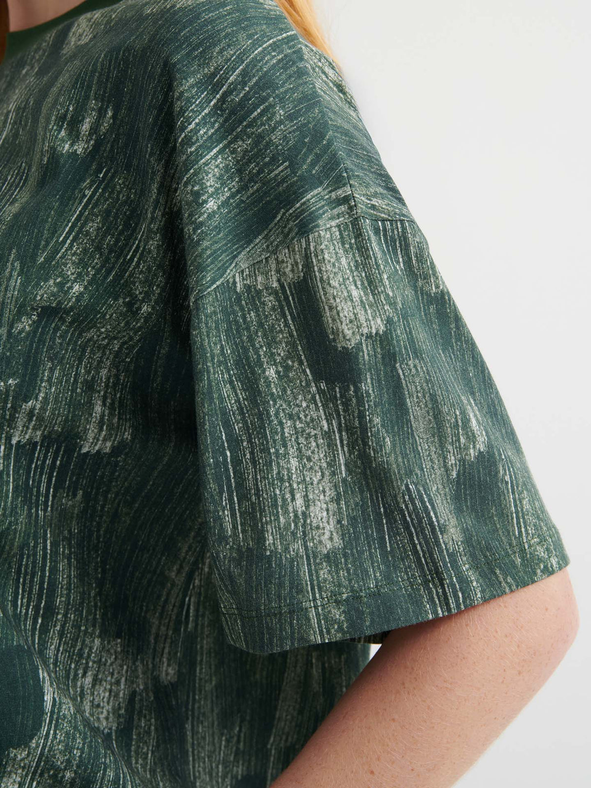 Close-up of a person wearing the Brushstroke Tee, an oversized boxy dark green shirt with a white abstract brushstroke pattern and drop shoulder sleeves.