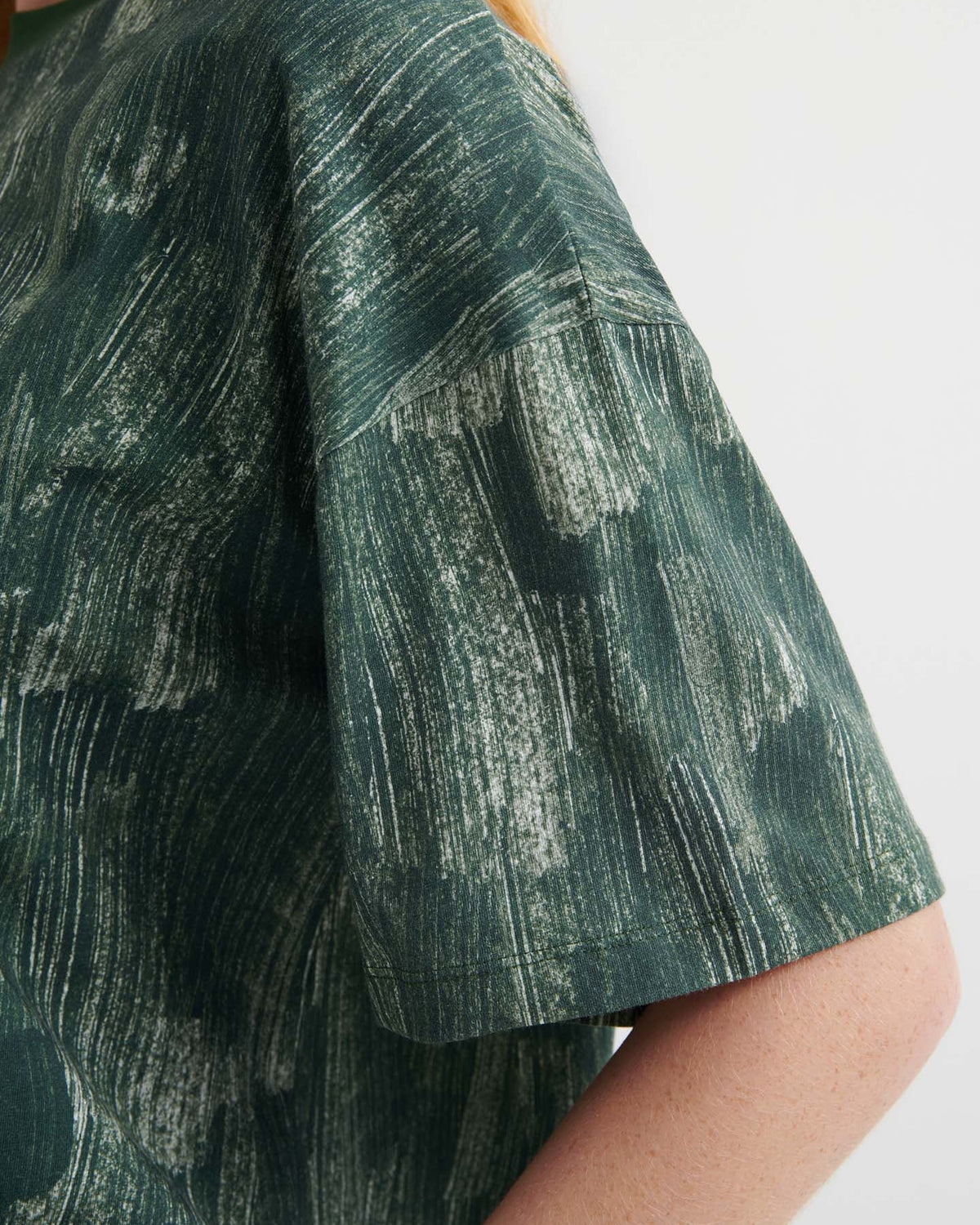 Close-up of a person wearing the Brushstroke Tee, an oversized boxy dark green shirt with a white abstract brushstroke pattern and drop shoulder sleeves.