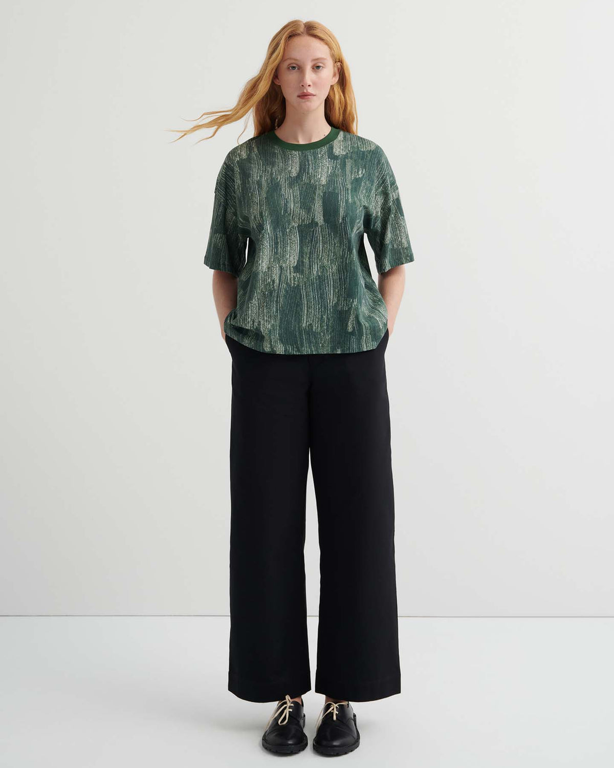 A person with long hair stands against a plain background, wearing the oversized Brushstroke Tee with drop shoulder sleeves in a green pattern, paired with black wide-leg pants and black shoes.