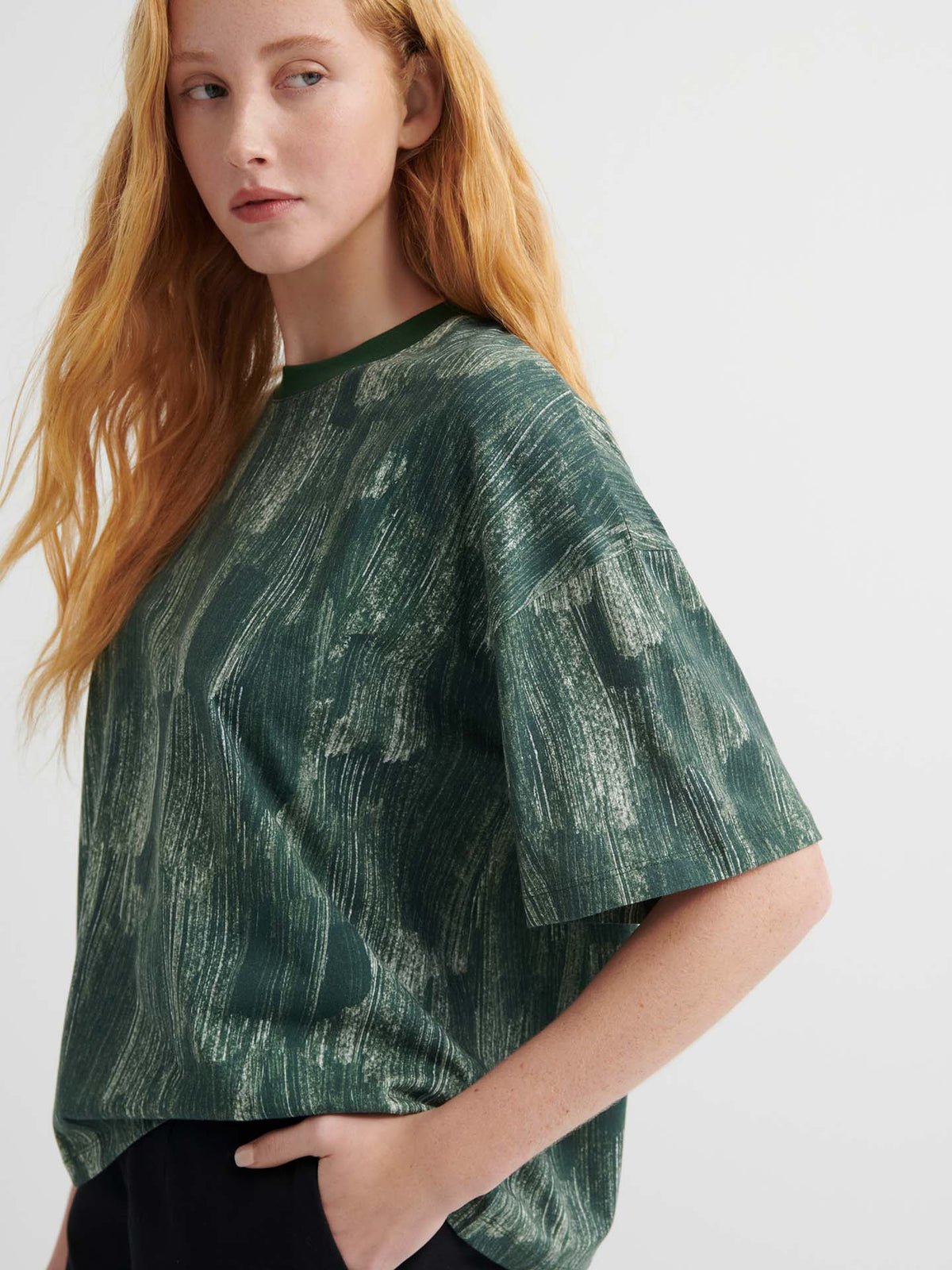 A person with long red hair wears the oversized Brushstroke Tee with a green pattern and drop shoulder sleeves, paired with black pants, standing against a plain light background.