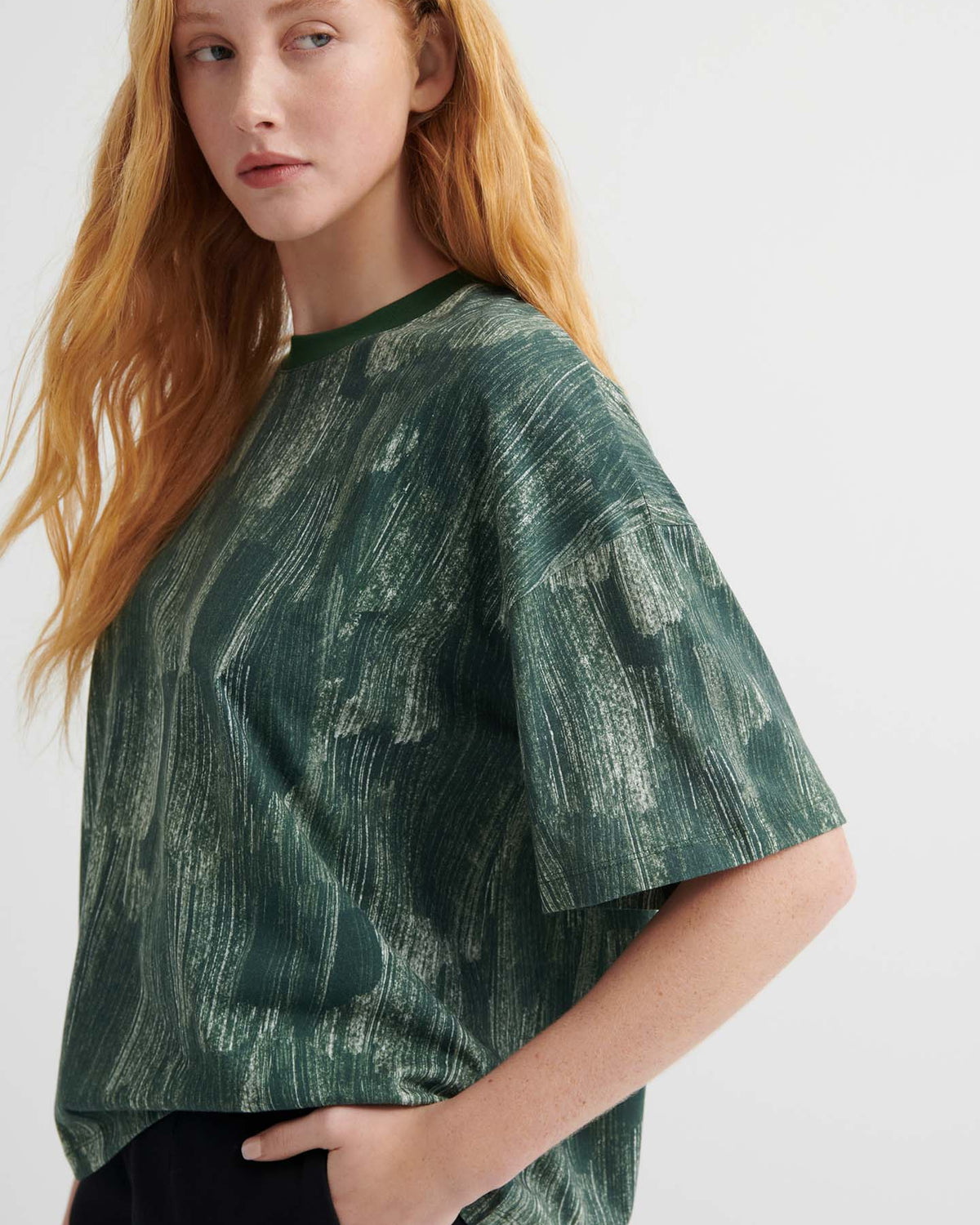 A person with long red hair wears the oversized Brushstroke Tee with a green pattern and drop shoulder sleeves, paired with black pants, standing against a plain light background.