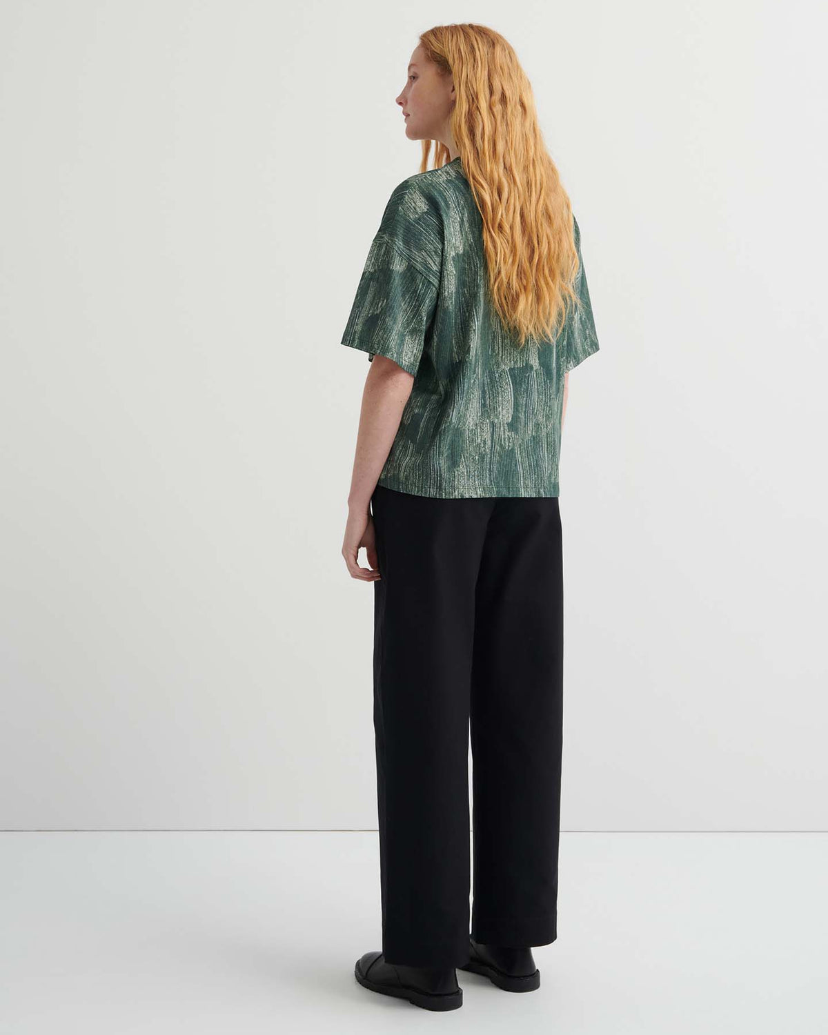 A person with long, wavy hair faces away, wearing a green Brushstroke Tee with drop shoulders, black wide-leg pants, and black shoes against a plain white background.