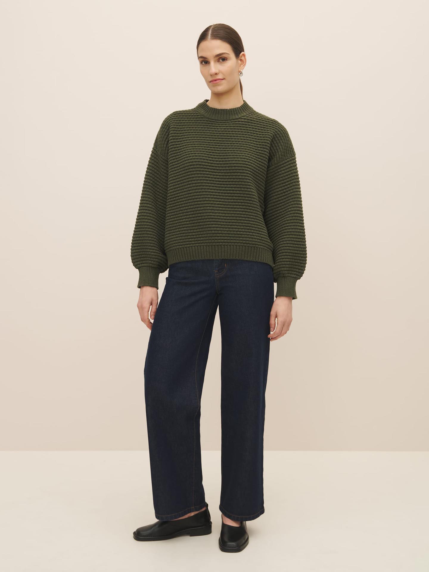 Woman standing against a neutral background, wearing a Kowtow Bubble Jumper in Khaki Marle and dark blue jeans with black loafers.
