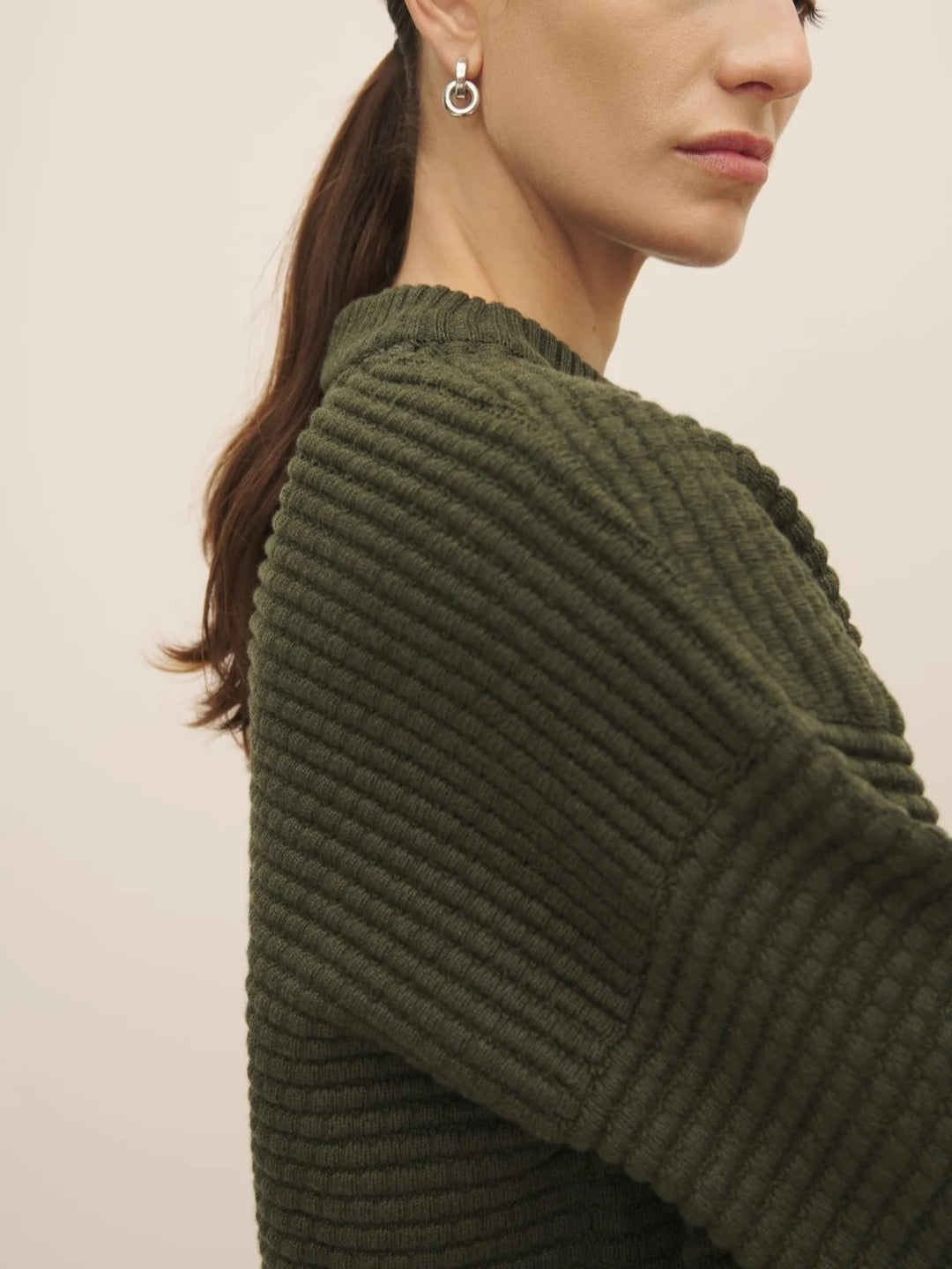 Profile view of a woman wearing a Kowtow Bubble Jumper – Khaki Marle, made from fairtrade organic cotton, and an earring with a neutral background.