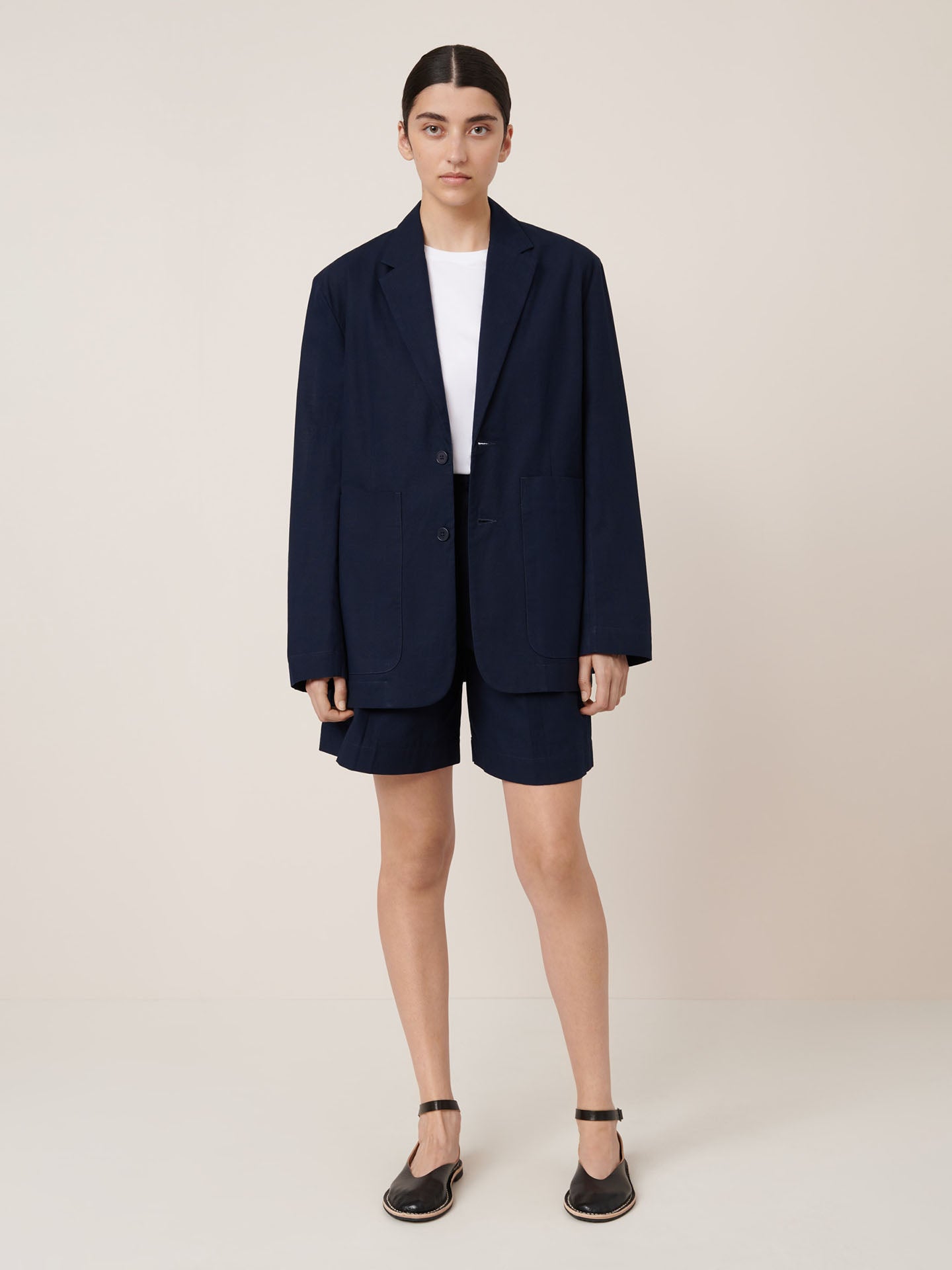 A person stands against a plain background wearing the Kowtow Carter Blazer in Evening Navy with matching shorts, a white t-shirt, and black sandals. Check our size guide for precise garment measurements.