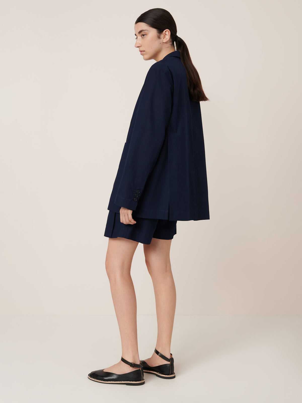 A person stands sideways against a plain background, wearing the Kowtow Carter Blazer in Evening Navy with matching shorts and black ankle-strap flats. For detailed garment measurements, please refer to our size guide.