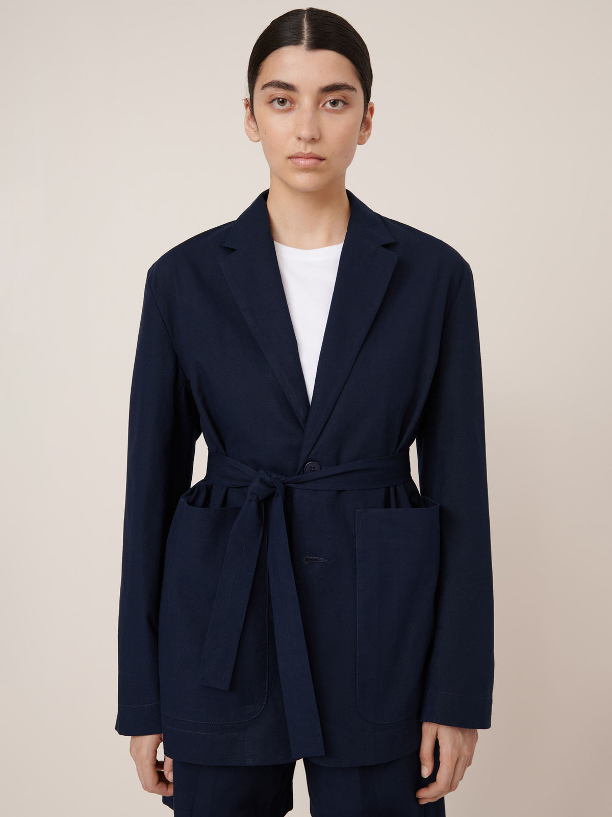 A person stands facing forward against a plain light background, wearing the Kowtow Carter Blazer in Evening Navy with an oversized fit and belted waist over a white shirt.