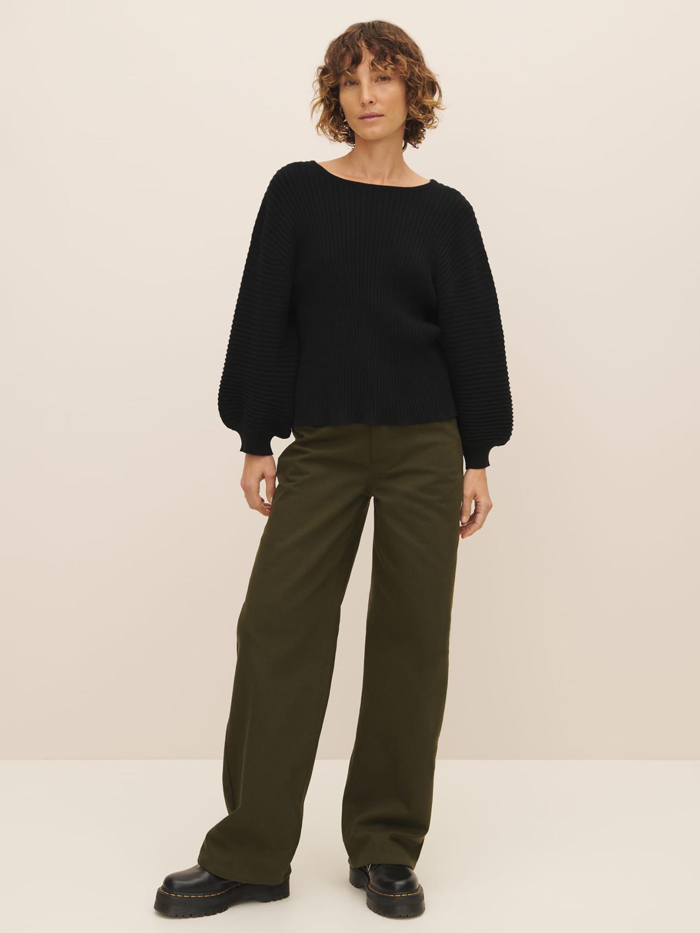 A woman stands against a neutral background wearing a black rib knit Cassia Sweater made of Fairtrade organic cotton and olive green trousers with black platform shoes from Kowtow.
