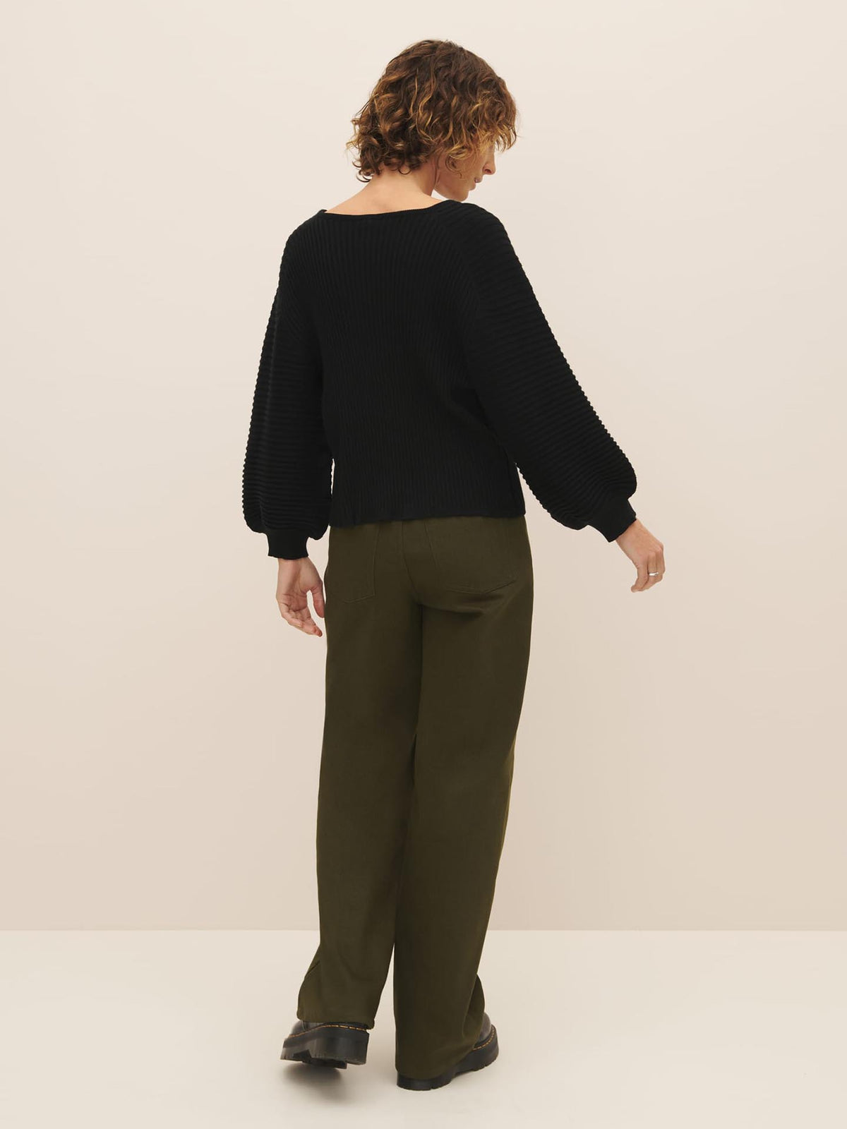 Woman standing with her back to the camera wearing a Kowtow Cassia Sweater in Black and green trousers.
