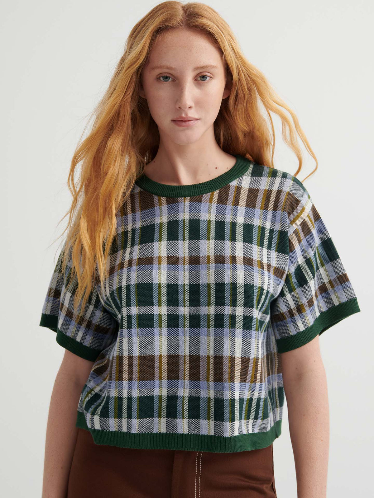 A person with long red hair wears the Chequered Tee – Forest, an oversized green and brown tee with drop shoulder sleeves, paired with brown pants against a plain light background.