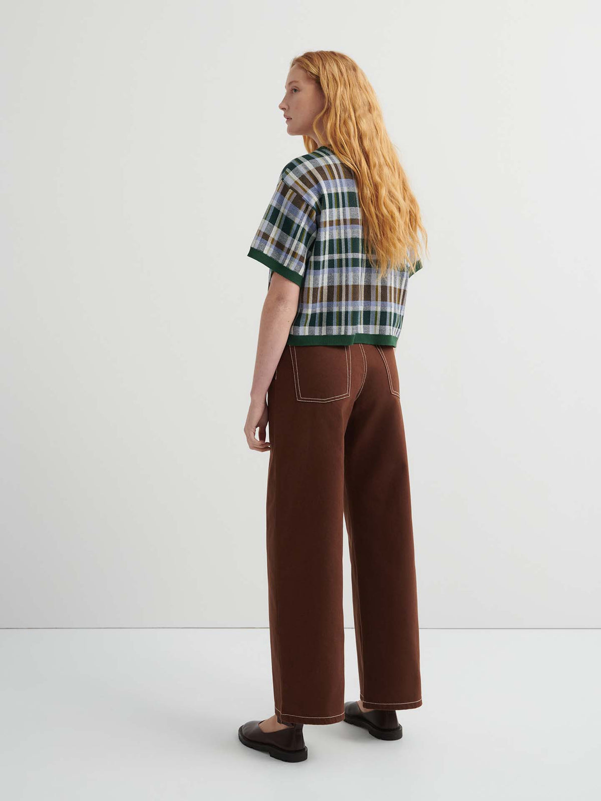 A person with long red hair faces away, wearing the Chequered Tee – Forest with drop shoulder sleeves, wide-legged brown pants with white stitching, and black shoes against a plain background.