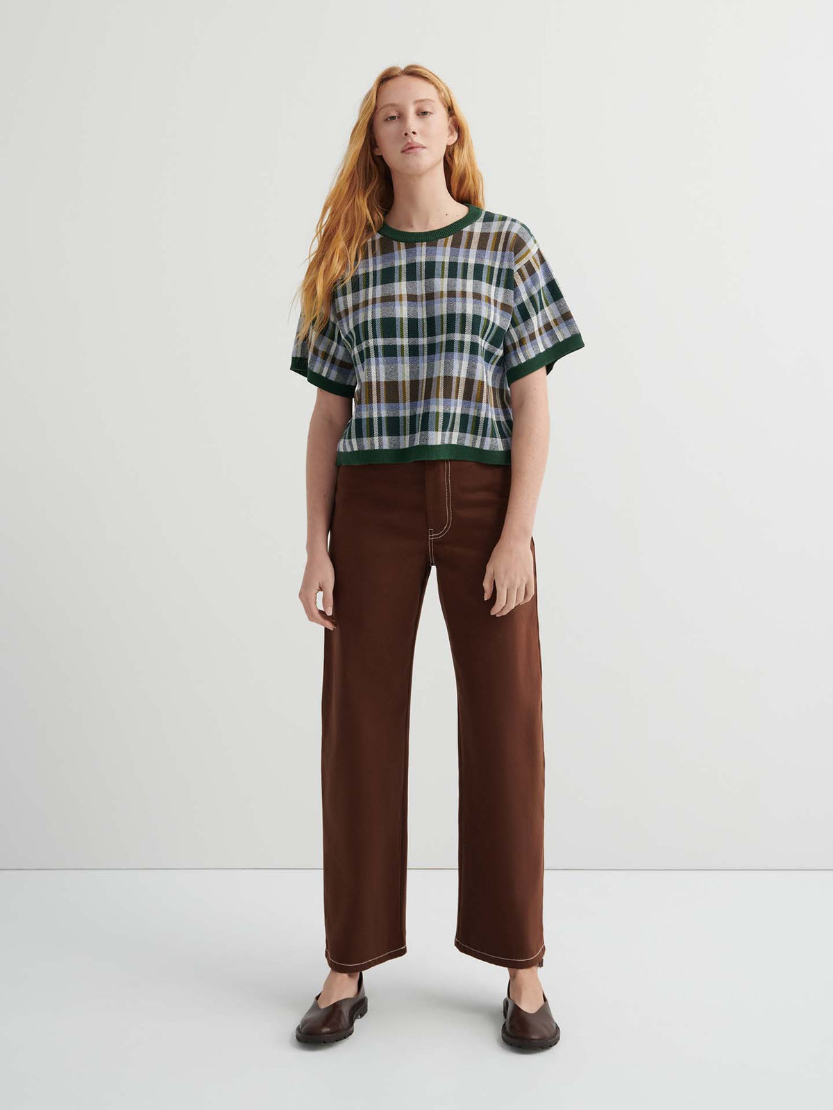 A person with long red hair wears the Chequered Tee – Forest with drop shoulder sleeves, wide-leg brown pants, and black shoes, standing against a plain white background.