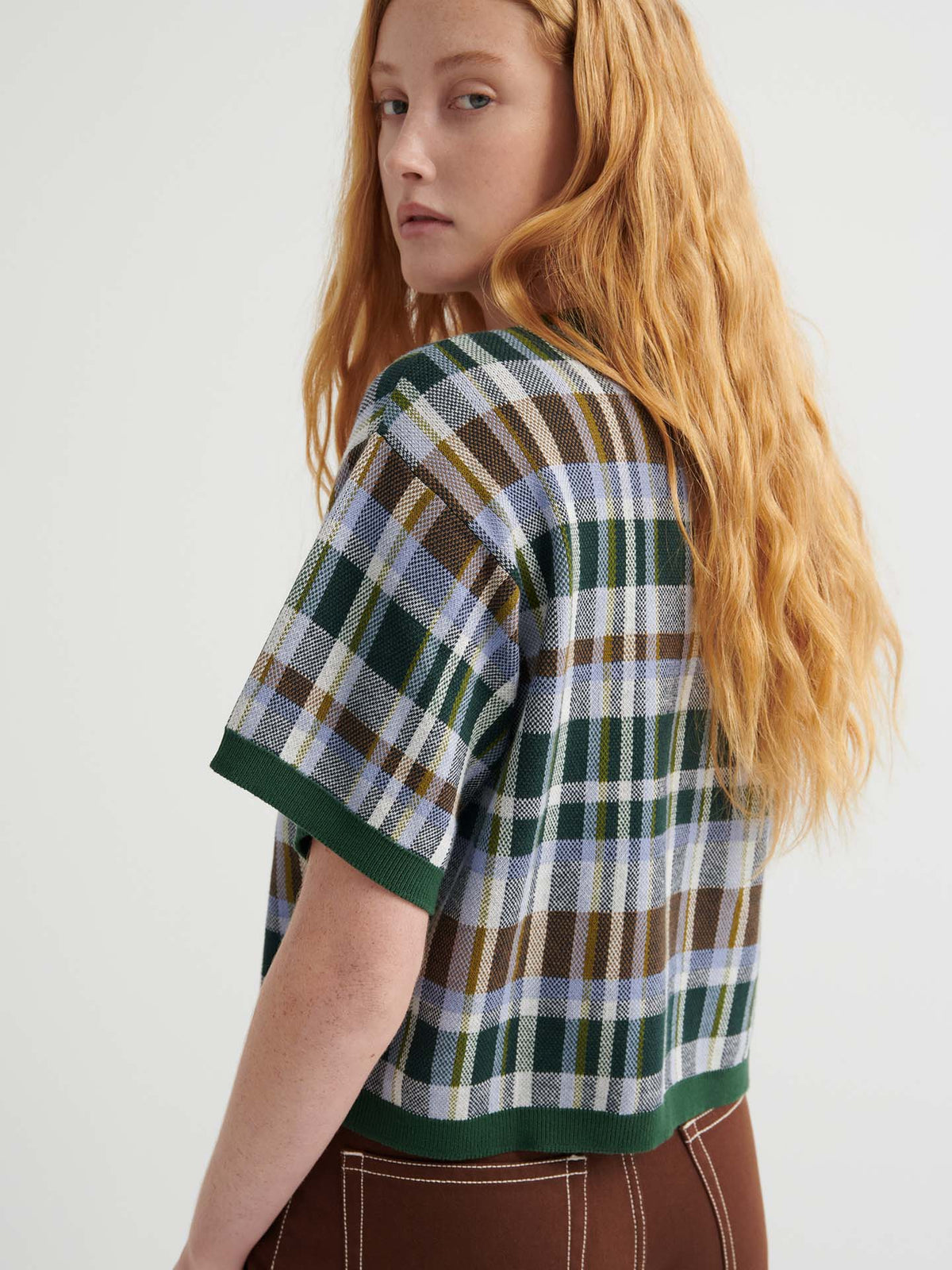 A person with long wavy red hair wears the Chequered Tee – Forest, featuring an oversized fit and drop shoulder sleeves in green, blue, and brown tones, paired with brown pants against a plain light background.