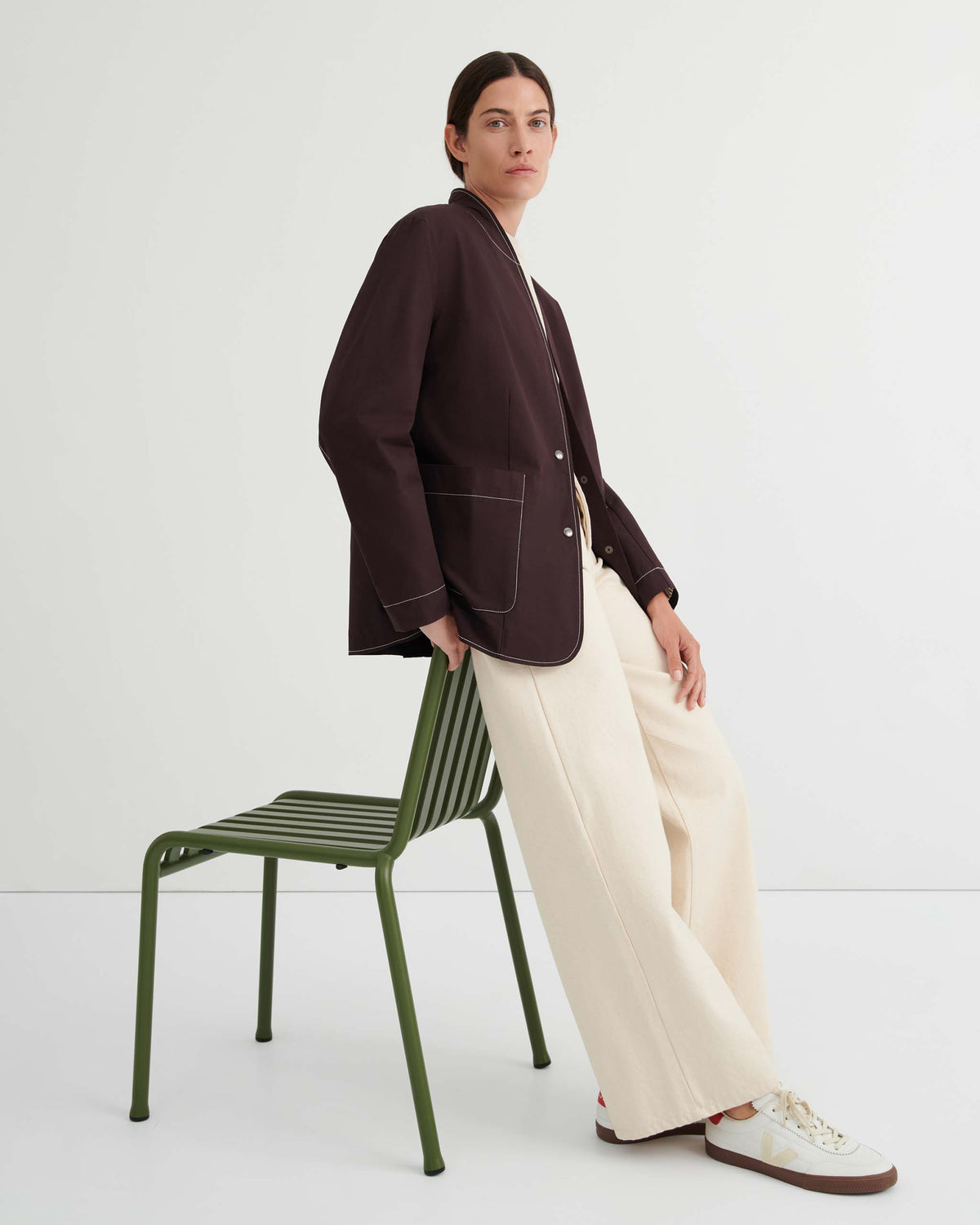 Person wearing a dark blazer, cream wide-leg pants, and sneakers sits sideways on a green metal chair against a plain white background.