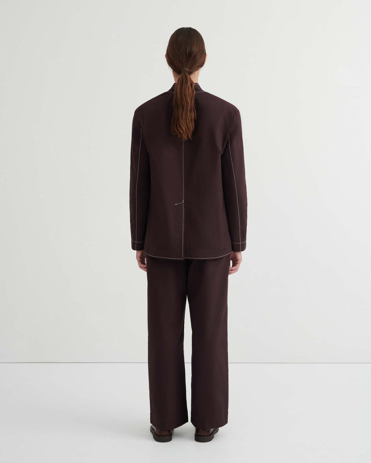 Person with long hair wearing a dark brown suit stands facing away from the camera against a plain white background.