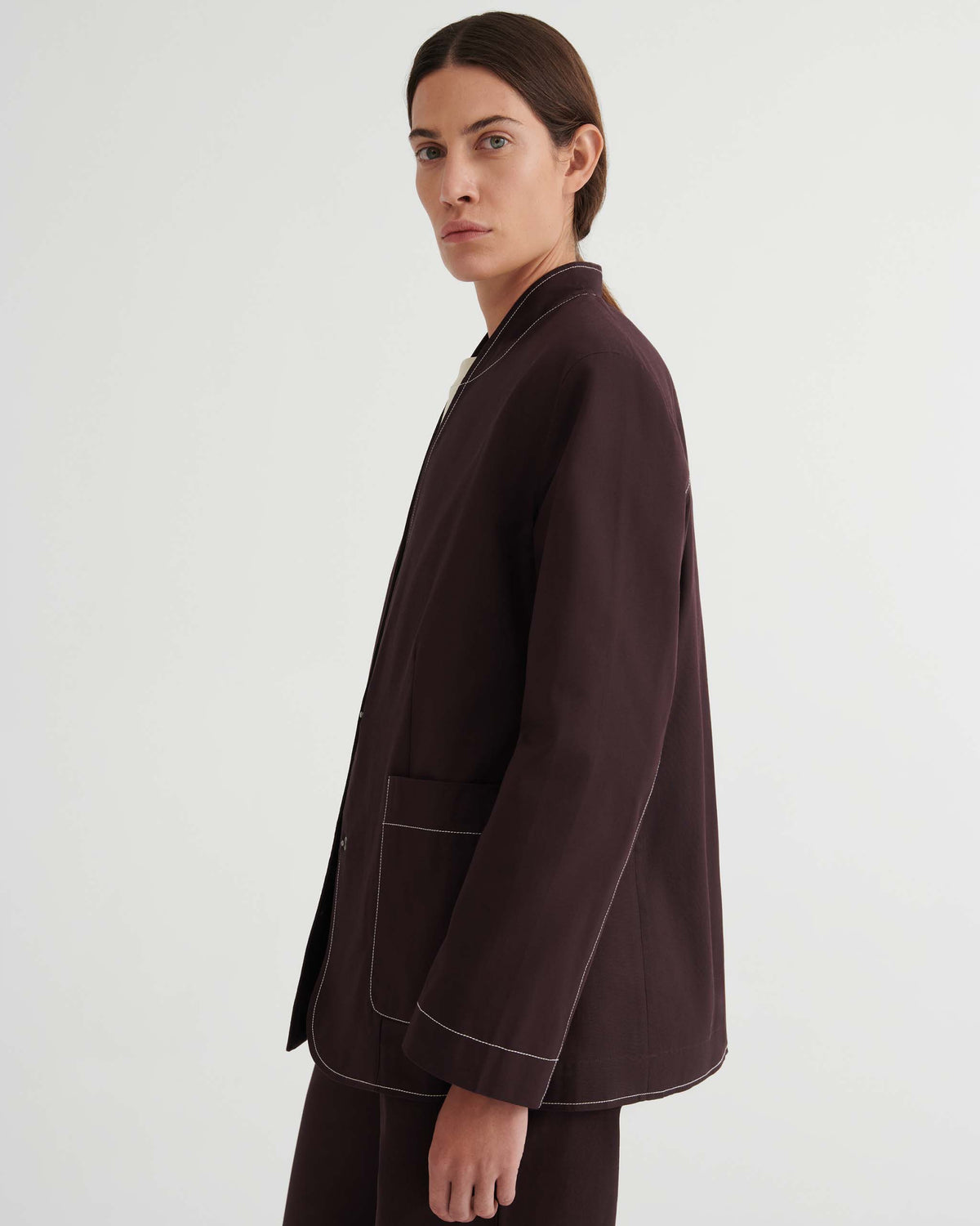 Person with long brown hair wears a dark brown jacket with white stitching, standing sideways and looking at the camera against a plain white background.