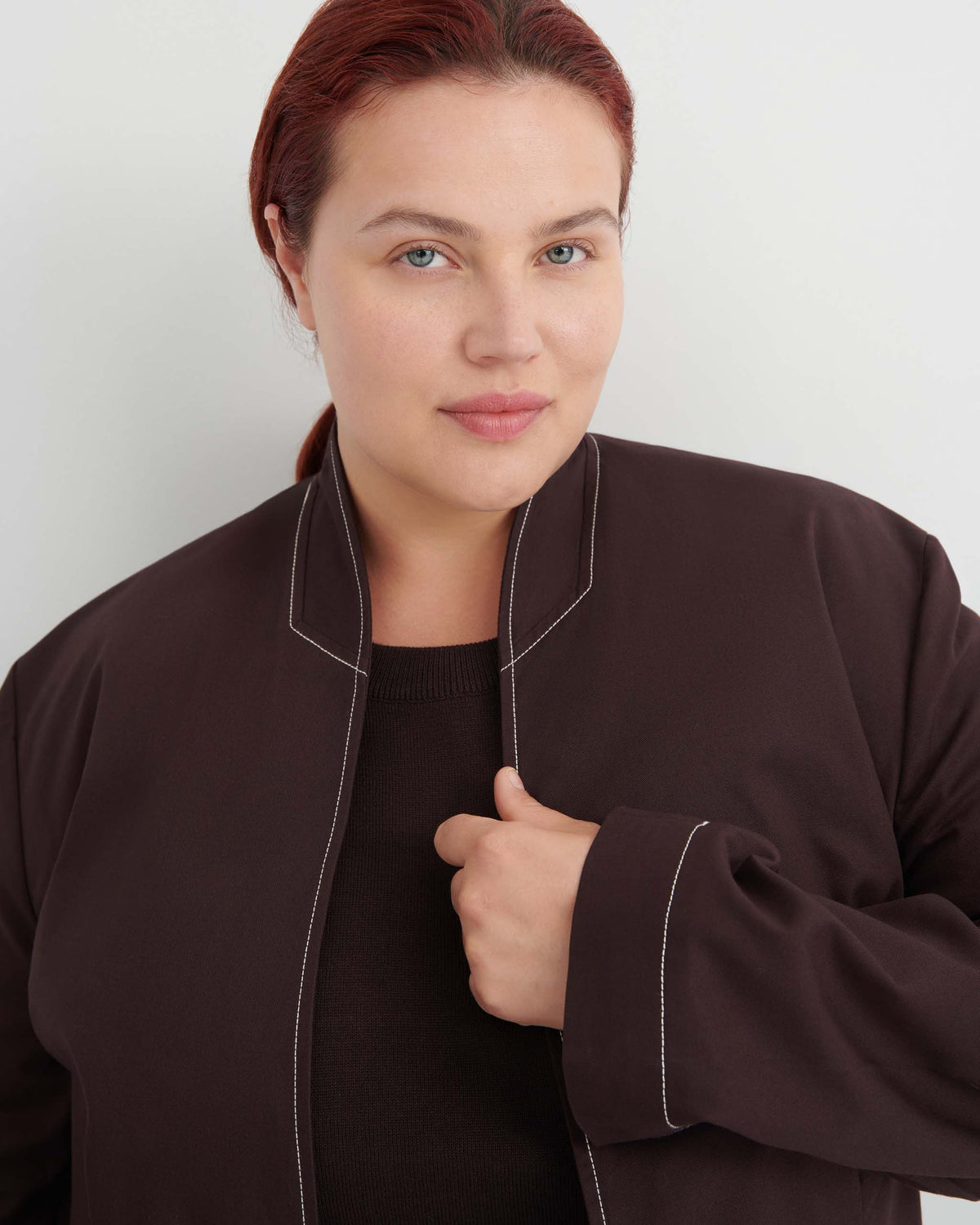 A person with light skin and red hair, wearing a dark brown jacket over a matching top, stands against a plain white background.