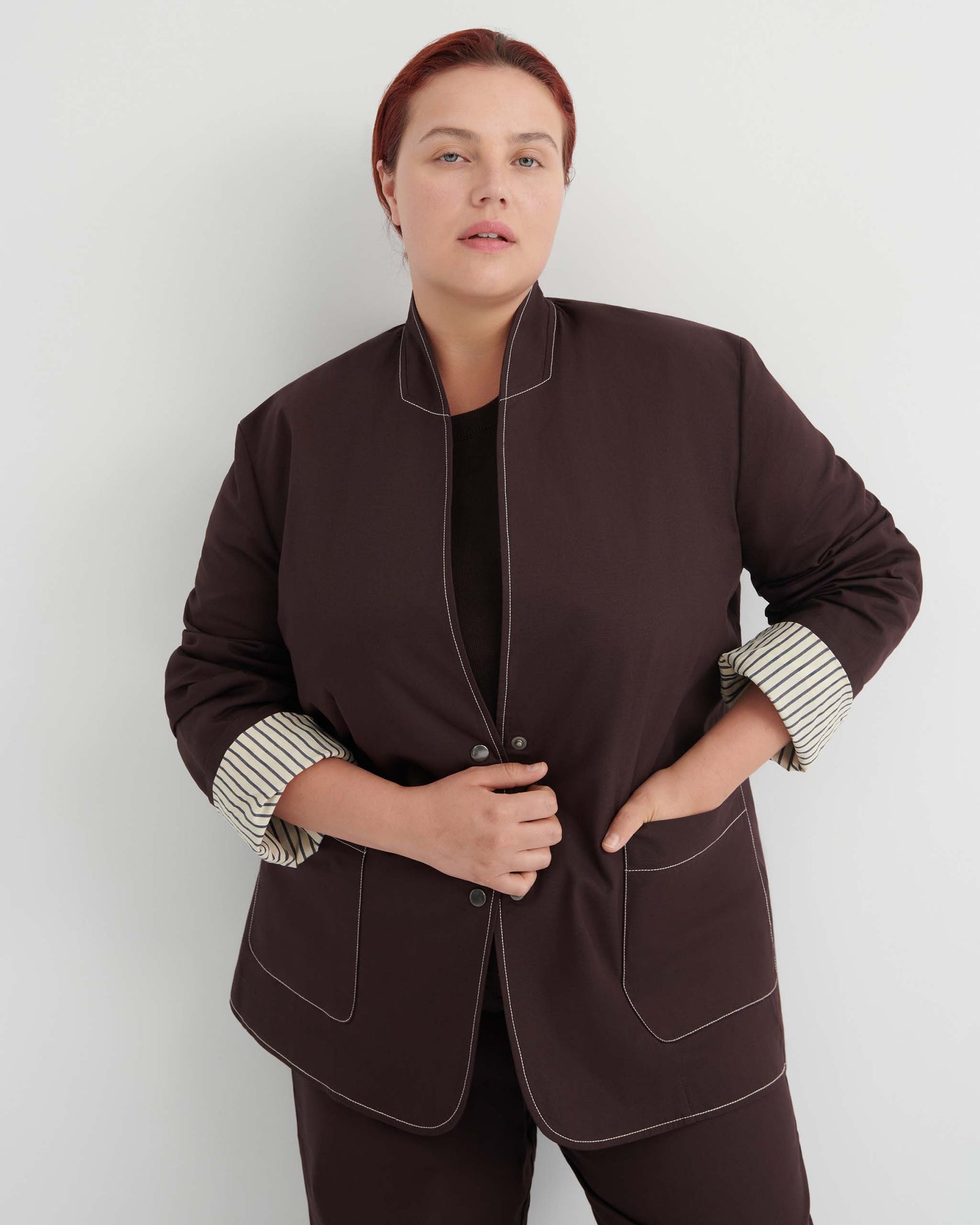 Person standing against a plain white background, wearing a dark jacket with striped cuffs and matching pants, looking directly at the camera with one hand holding the jacket closed.