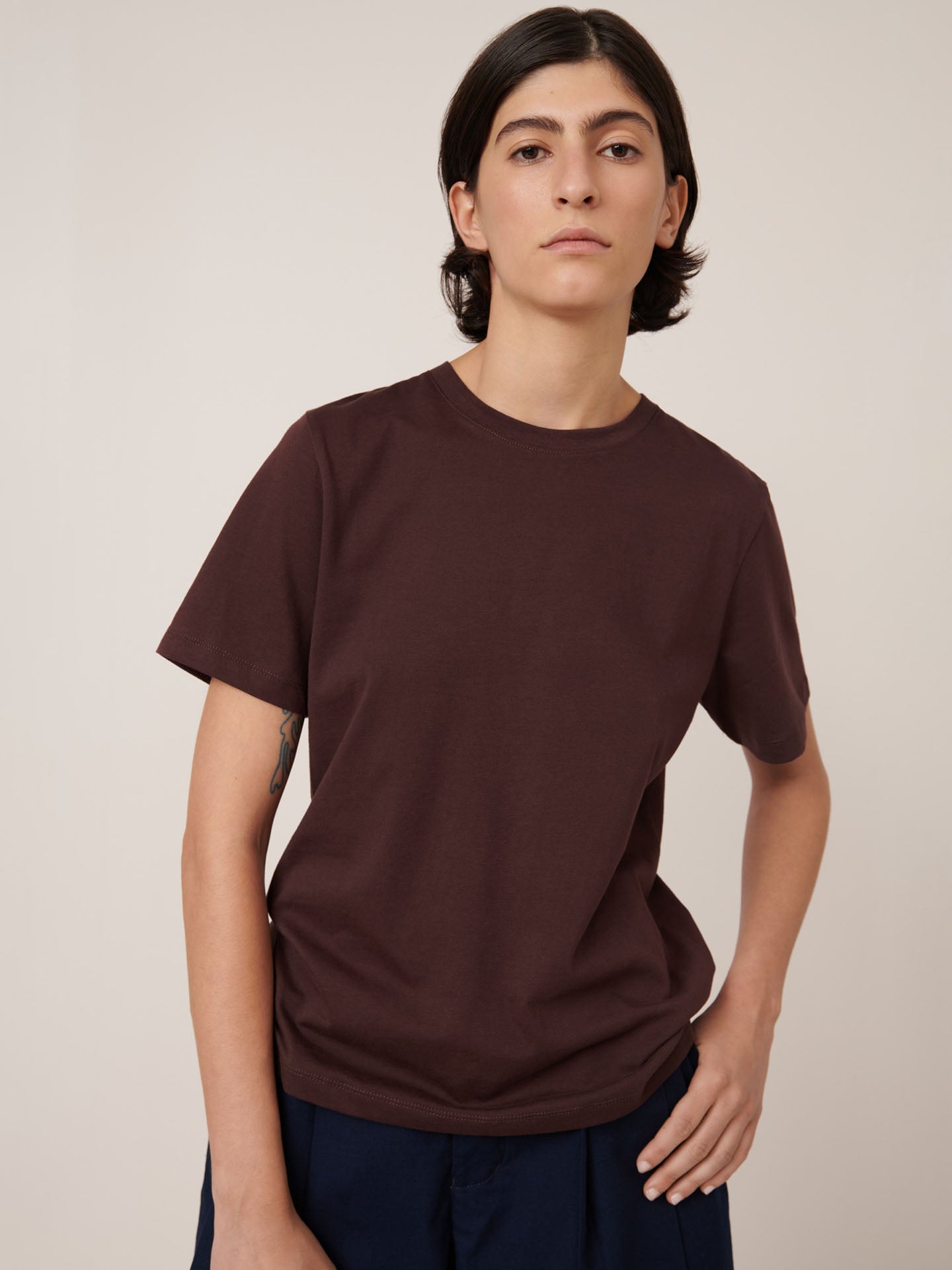 A person with straight, dark hair wears the Kowtow Classic Tee in Mahogany, styled in a standard fit with dark pants, standing against a neutral backdrop.