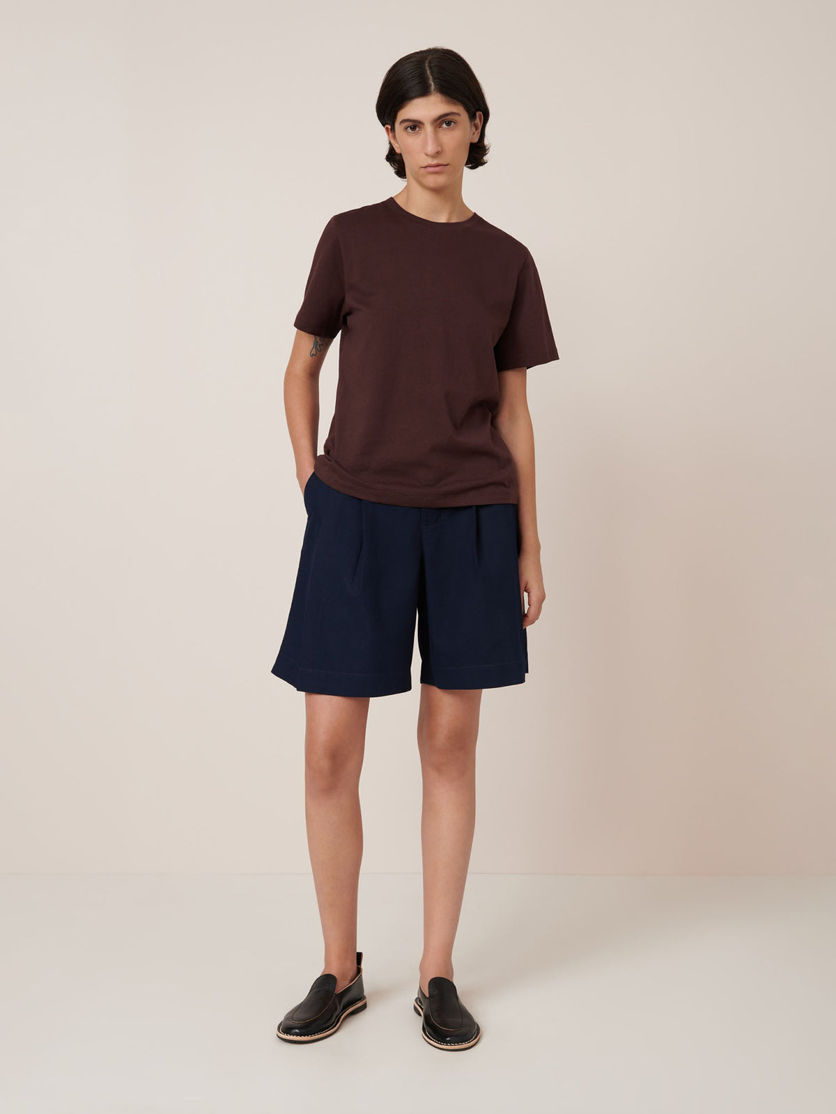 A person stands against a plain background wearing the Kowtow Classic Tee in Mahogany, navy shorts, and black slip-on shoes.
