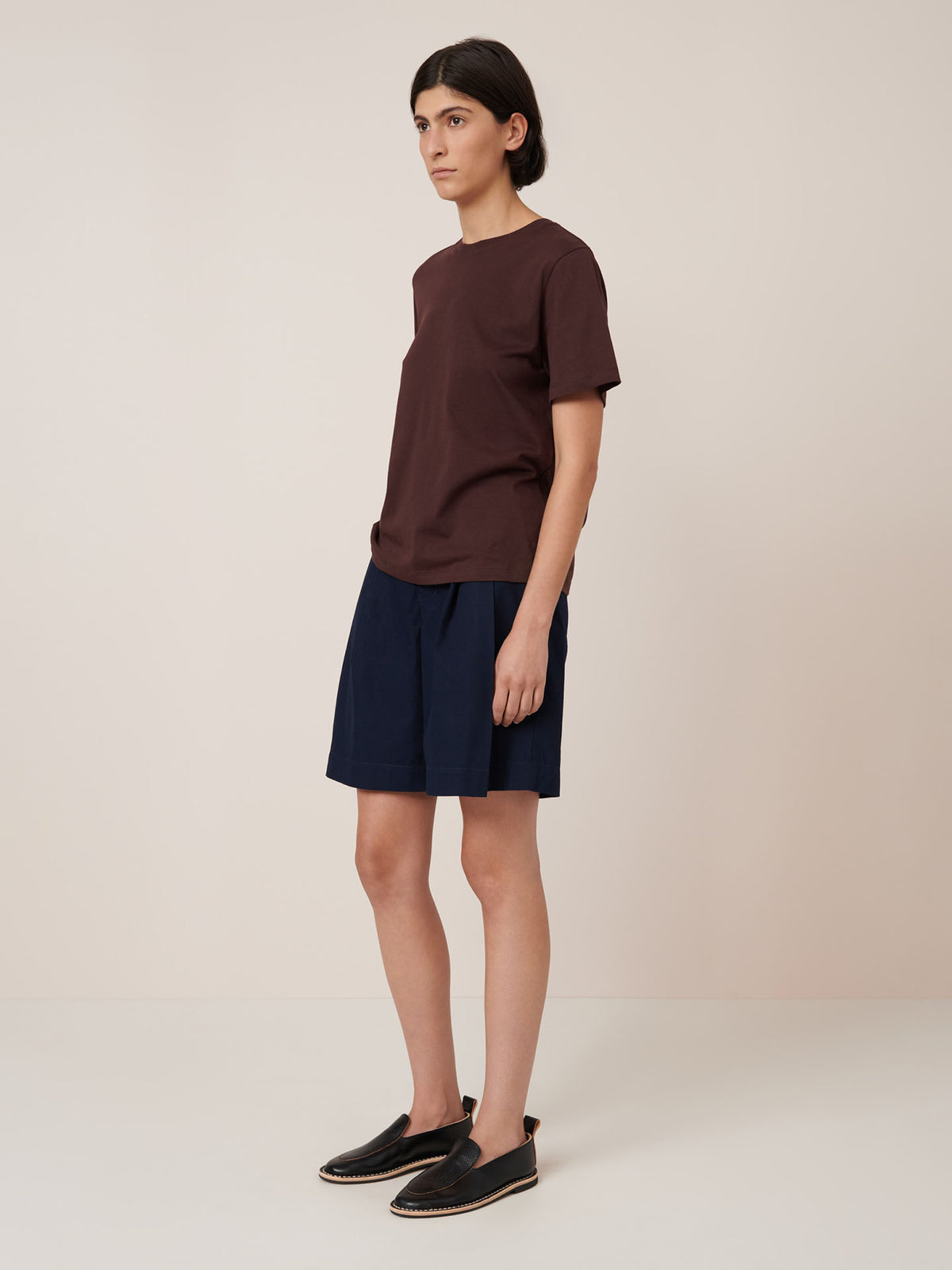 A person stands in a neutral room wearing the Kowtow Classic Tee in Mahogany, navy shorts, and black slip-on shoes, showcasing the standard fit based on Kowtow size garment measurements.