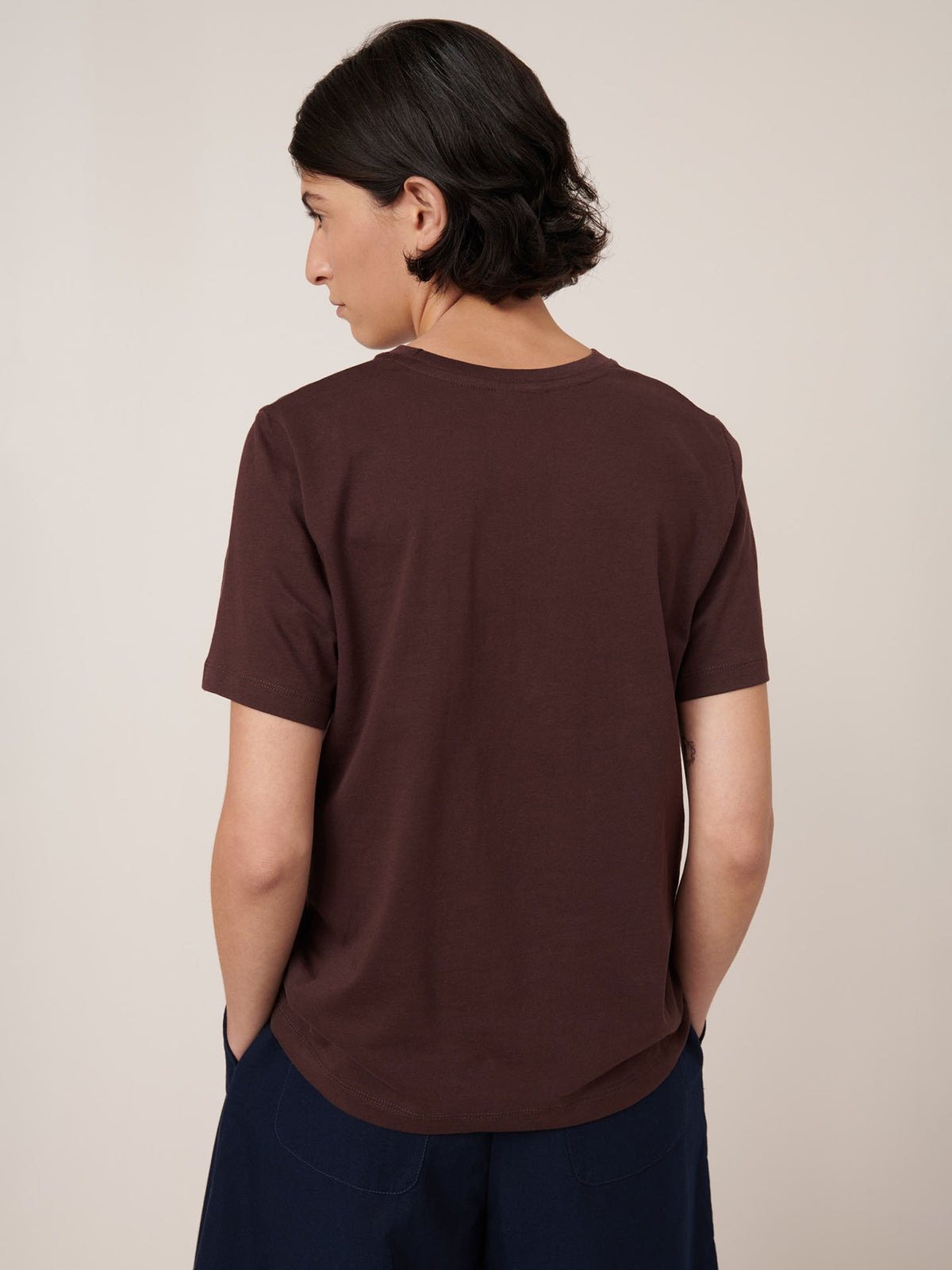 A person with short dark hair stands with their back to the camera, wearing the Kowtow Classic Tee in Mahogany and dark pants against a plain light background. Garment sizing follows Kowtow&#39;s standard guidelines.