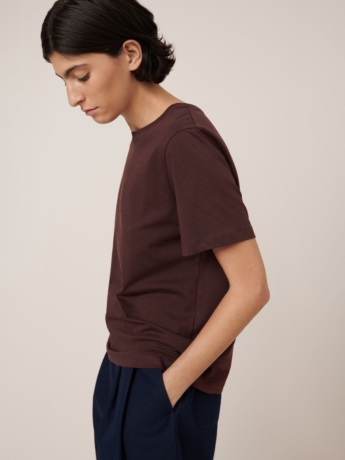 A person with short dark hair wears the Kowtow Classic Tee in Mahogany (standard fit) and navy pants, standing in profile with hands in pockets against a plain light background.