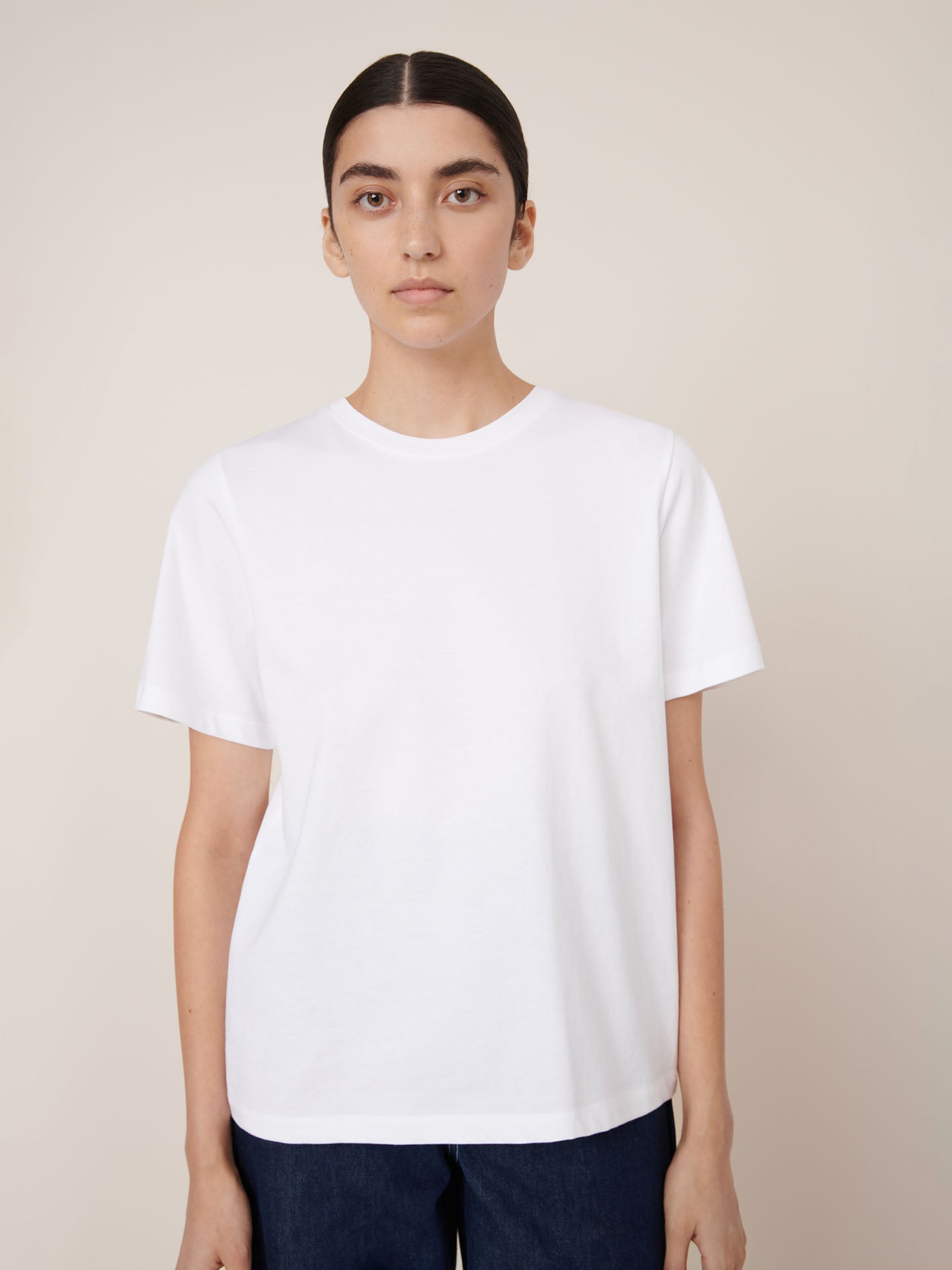 A person stands against a plain background, facing forward with a neutral expression, wearing the Kowtow Classic Tee – White in a relaxed fit paired with dark pants. The garment offers a comfortable, contemporary silhouette.