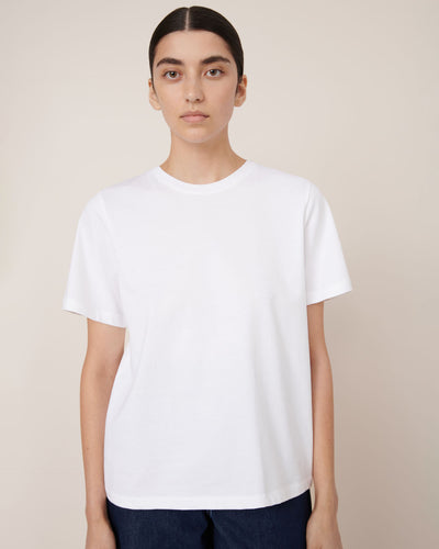 A person stands against a plain background, facing forward with a neutral expression, wearing the Kowtow Classic Tee – White in a relaxed fit paired with dark pants. The garment offers a comfortable, contemporary silhouette.