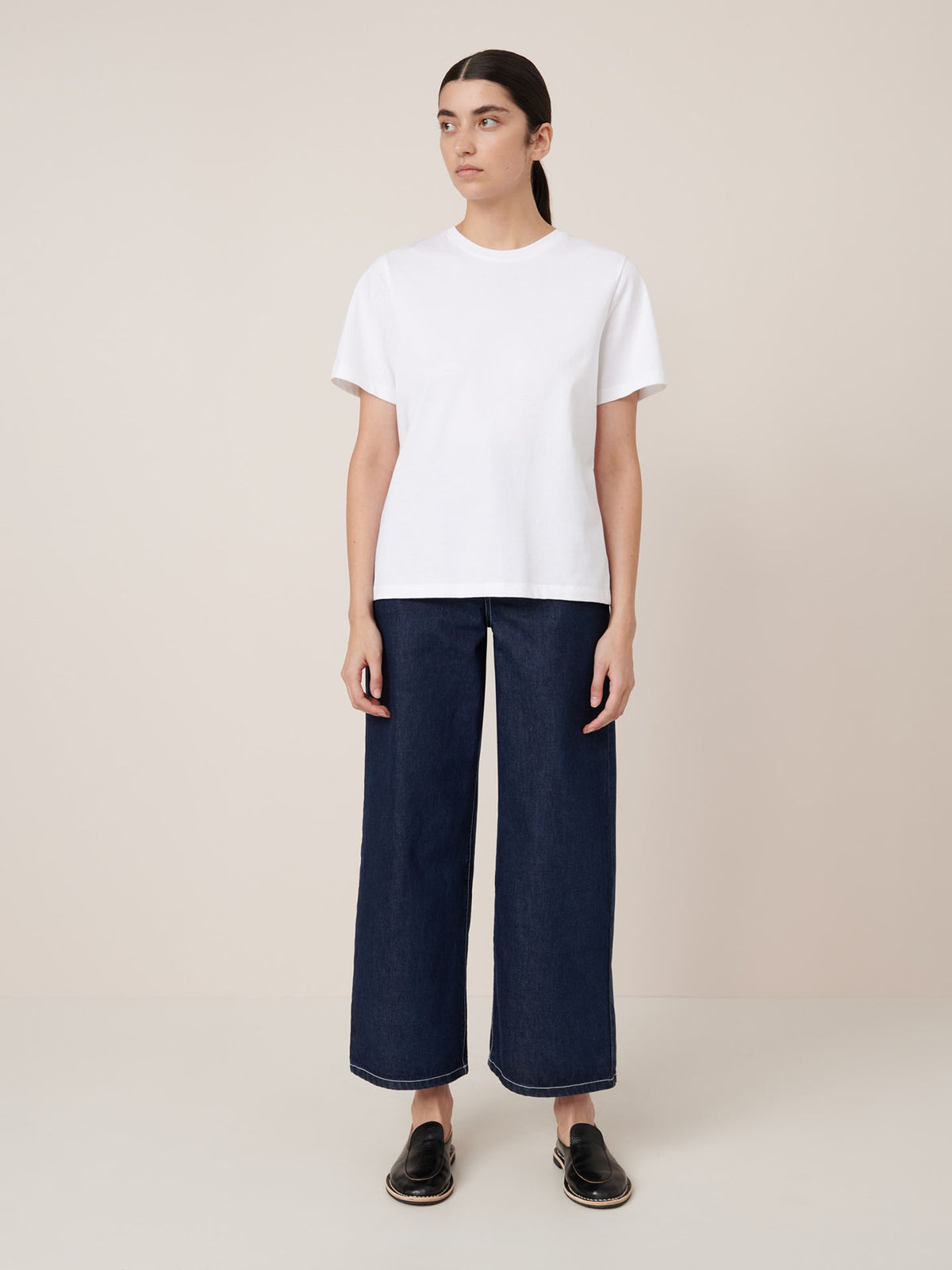 A person stands against a plain beige background wearing the Kowtow Classic Tee in white, paired with wide-leg dark blue jeans and black slip-on shoes.
