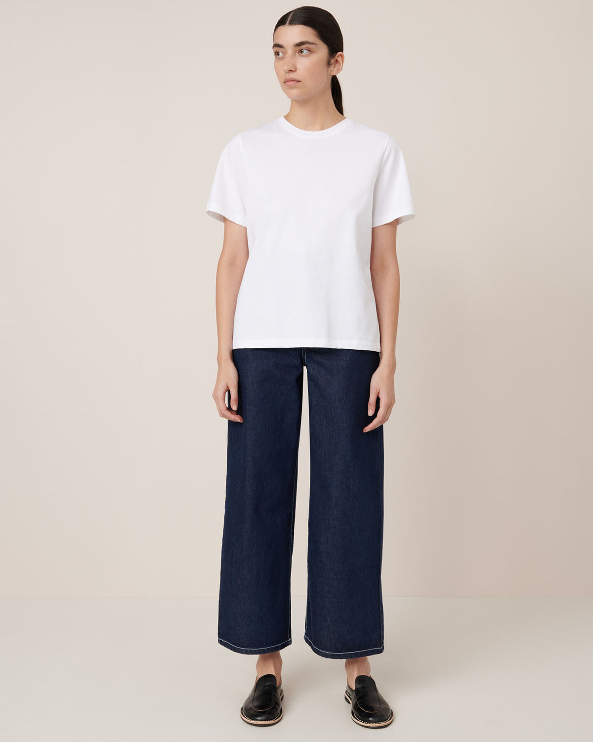 A person stands against a plain beige background wearing the Kowtow Classic Tee in white, paired with wide-leg dark blue jeans and black slip-on shoes.