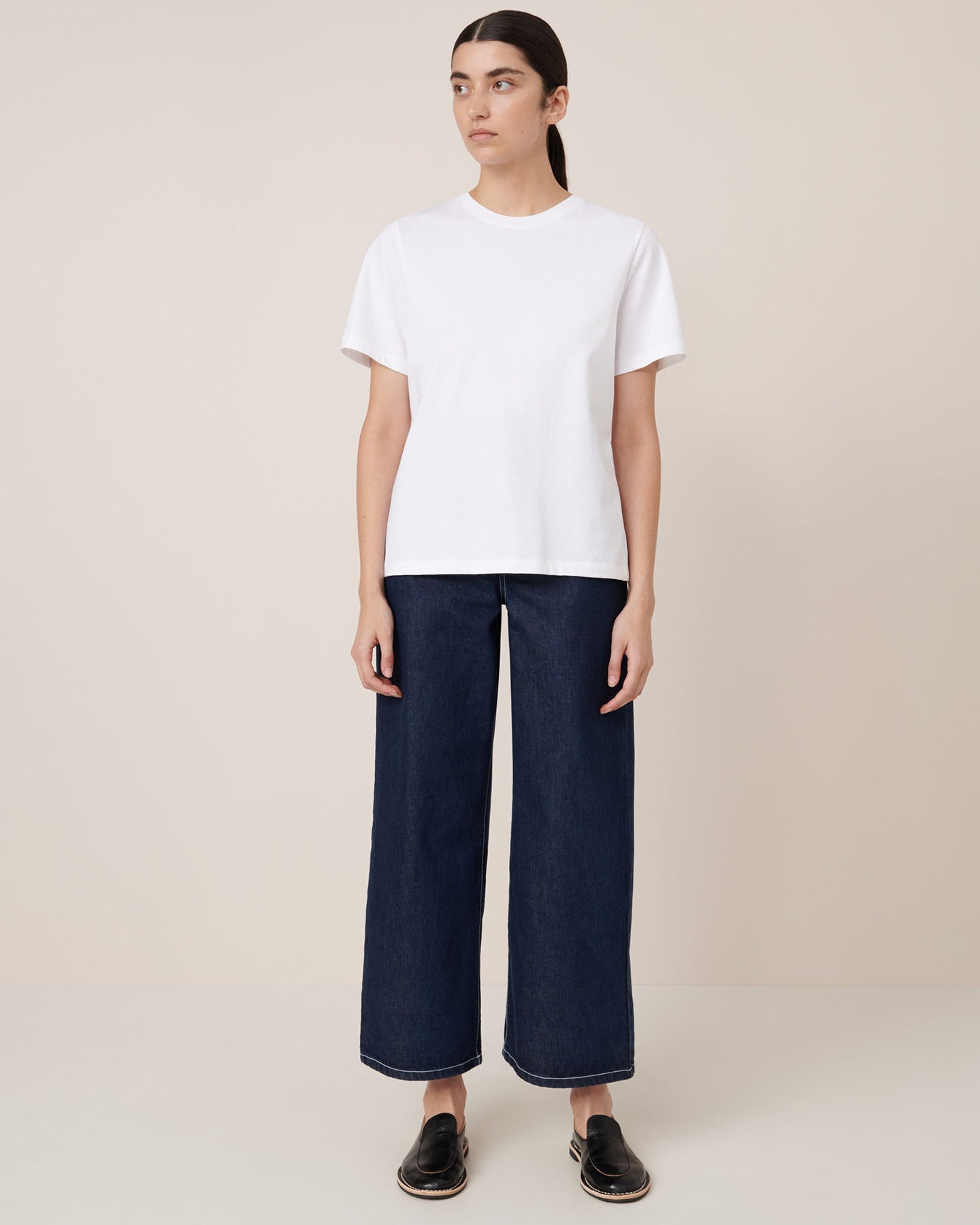 A person stands against a plain background, facing forward with a neutral expression, wearing the Kowtow Classic Tee – White in a relaxed fit paired with dark pants. The garment offers a comfortable, contemporary silhouette.