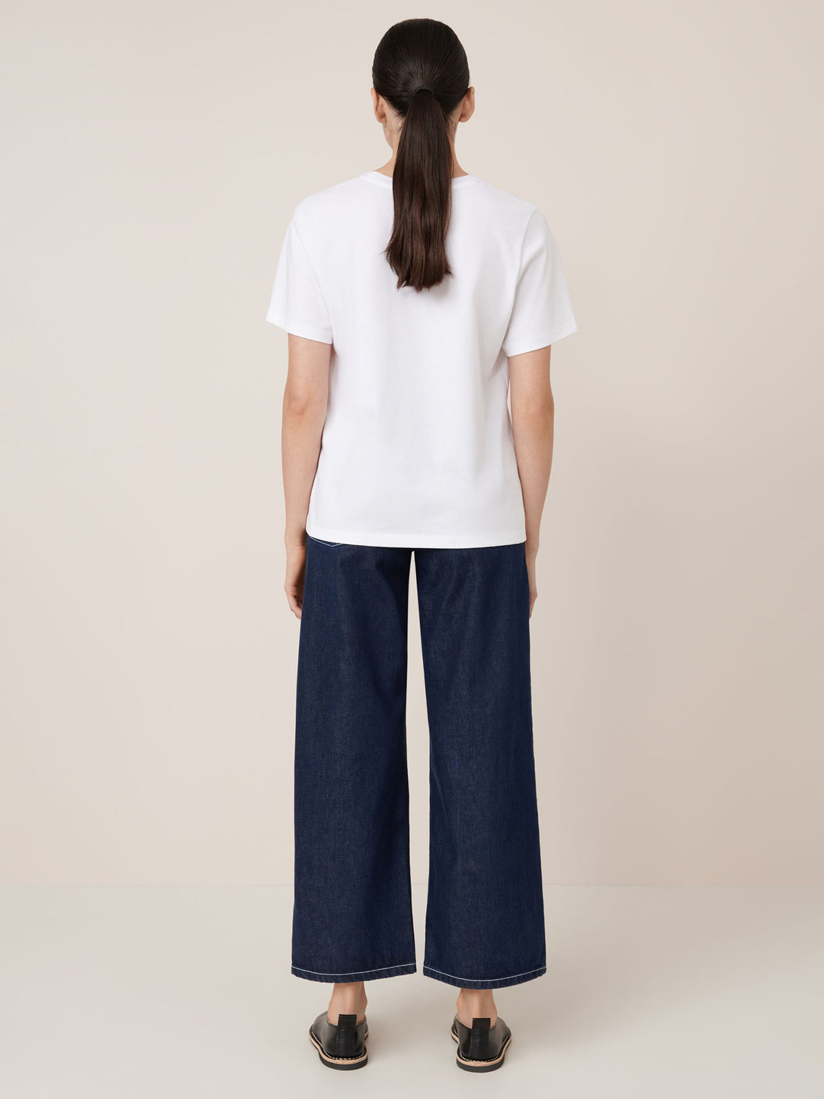 A woman faces away from the camera wearing Kowtow&#39;s Classic Tee in white, styled with wide-leg dark blue jeans and black slip-on shoes.