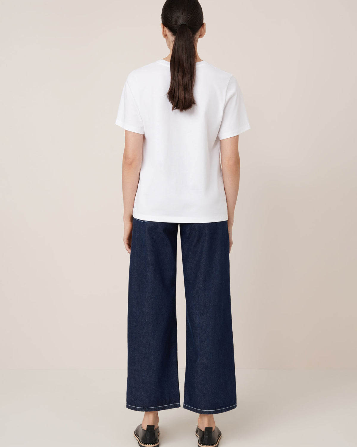 A woman faces away from the camera wearing Kowtow's Classic Tee in white, styled with wide-leg dark blue jeans and black slip-on shoes.
