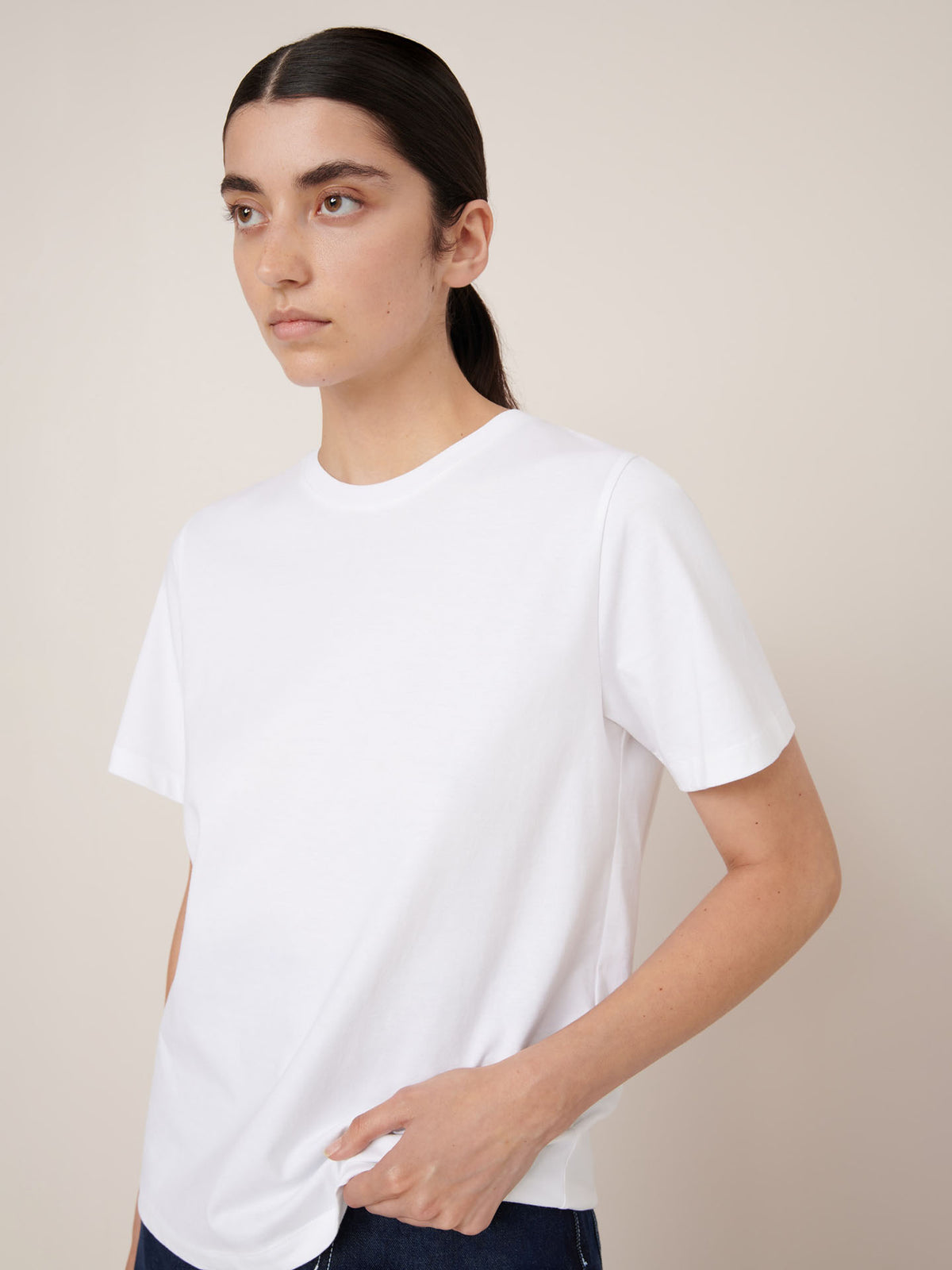 A person with long dark hair in a low ponytail wears the Kowtow Classic Tee – White, a relaxed fit short-sleeve t-shirt, standing against a light beige background.