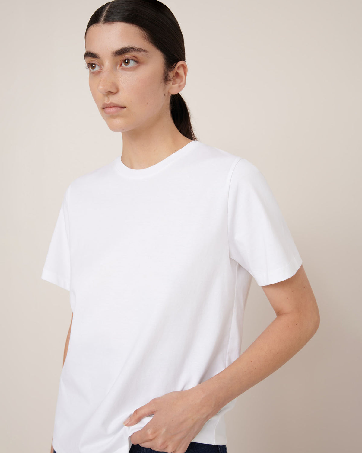 A person with long dark hair in a low ponytail wears the Kowtow Classic Tee – White, a relaxed fit short-sleeve t-shirt, standing against a light beige background.